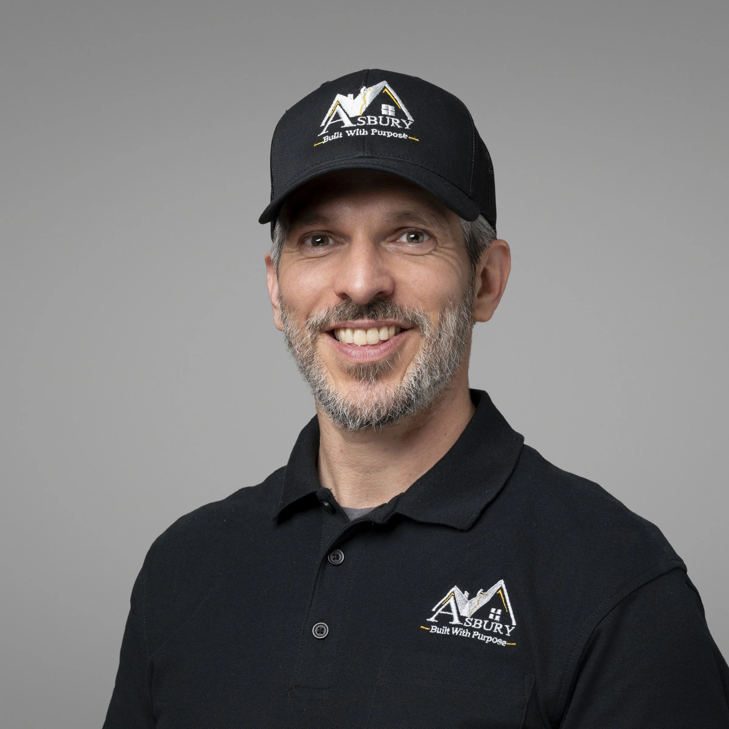Close-up of a man wearing a black baseball cap and black shirt, both with the logo and text 'ASBURY Built With Purpose' embroidered in white and yellow.