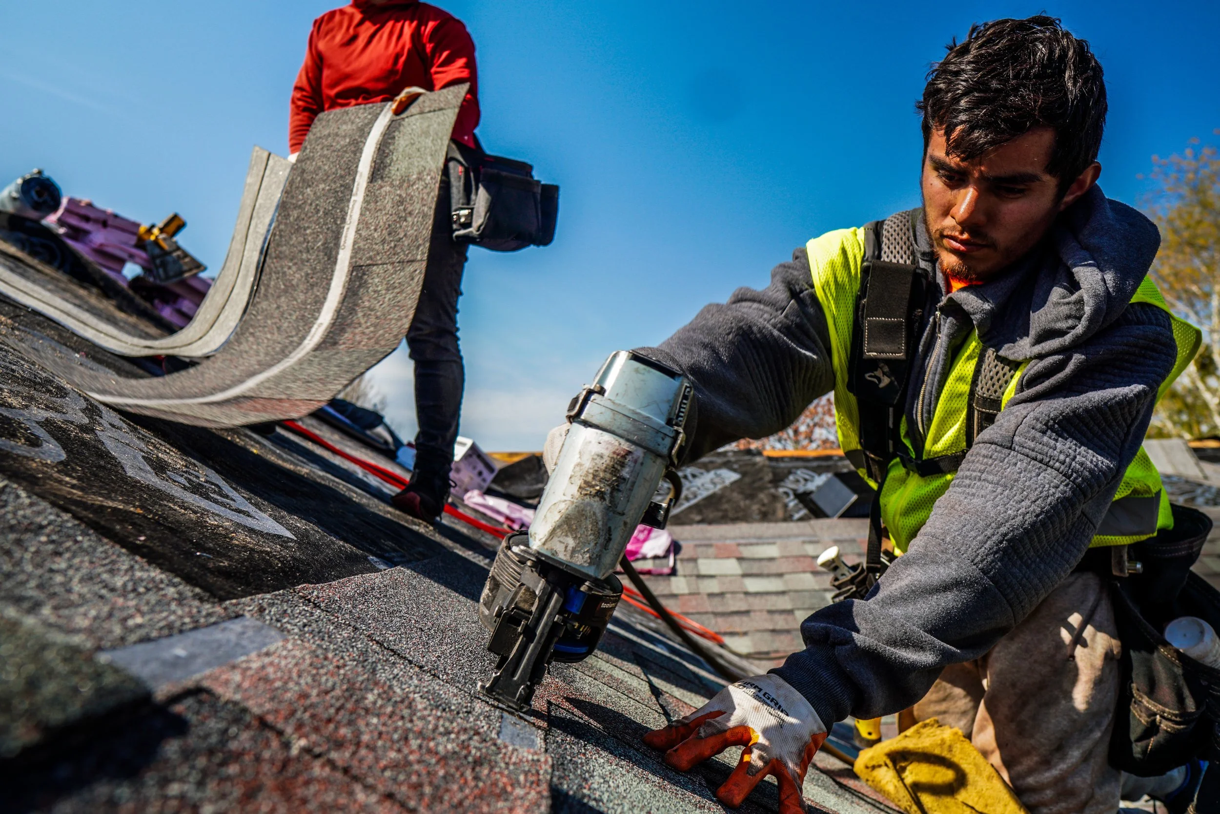 Two workers installing or repairing shingles on a roof, with one using a pneumatic nail gun and the other in the background handling tools, under a clear blue sky.