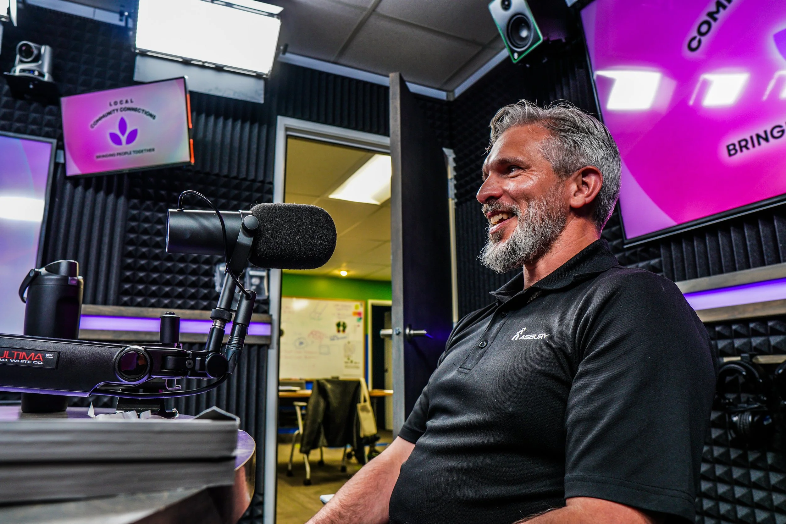 A man with gray hair and a beard smiling in a recording studio, sitting in front of a professional microphone with soundproofing foam on the walls, and multiple screens displaying graphics in purple and black colors.