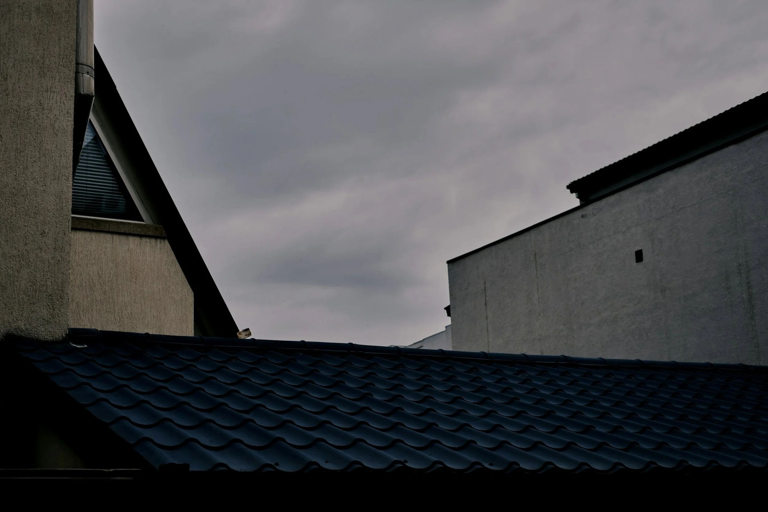 Overcast sky above rooftops of neighboring houses, one with a window and the other with a flat roof, with a tiled roof in the foreground.