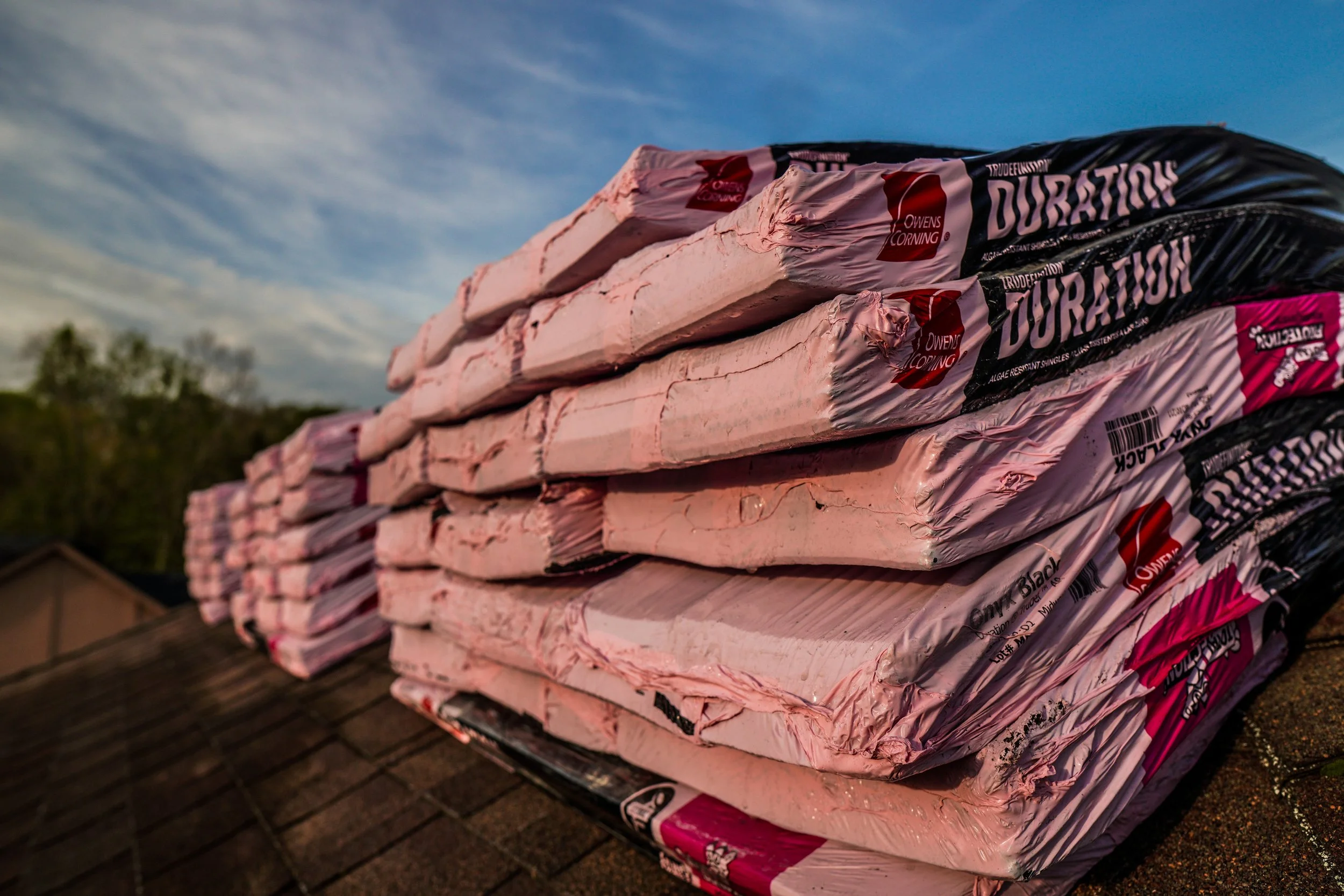 Stacked bags of Owens Corning pink fiberglass insulation on a wooden surface outdoors with a partly cloudy sky in the background.