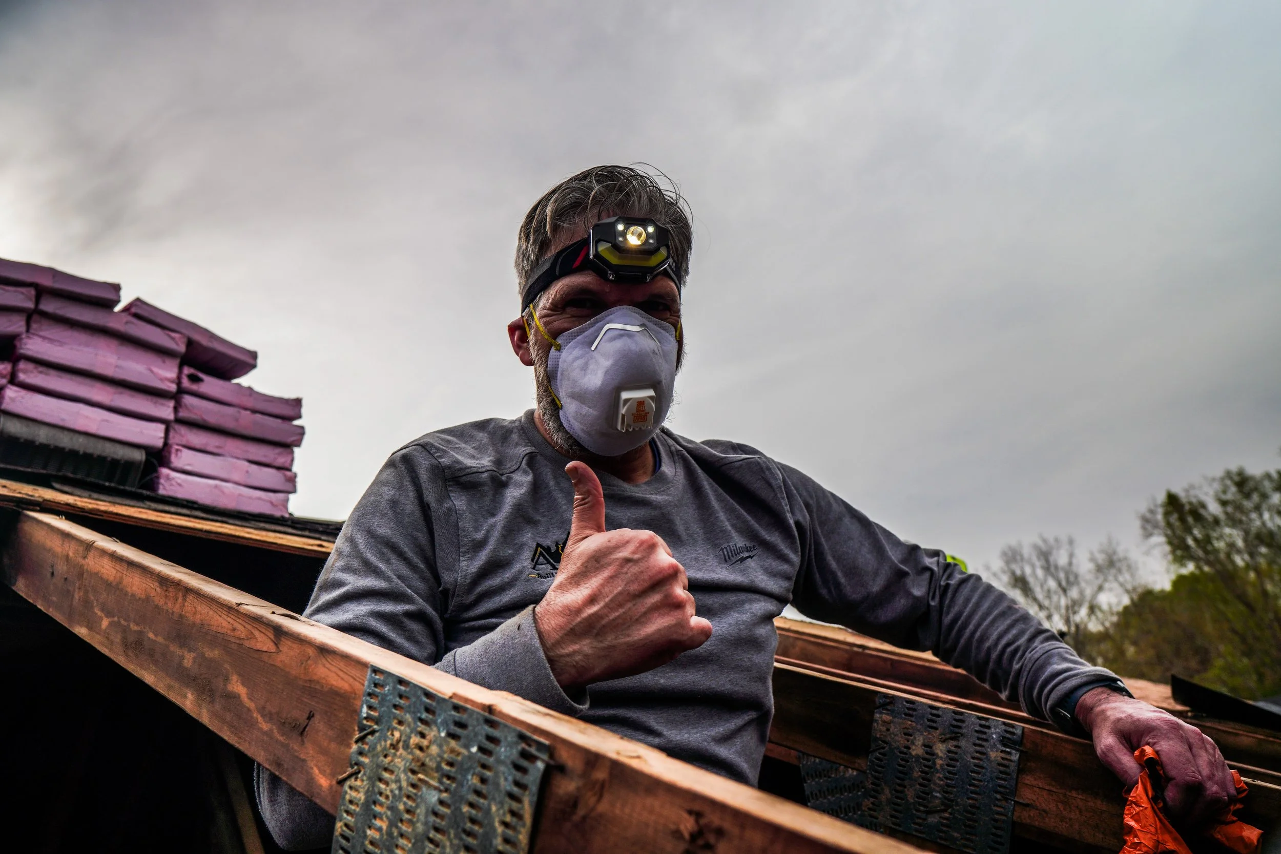 Man wearing a headlamp and a face mask giving a thumbs up on a construction site with a wooden frame and purple insulation foam