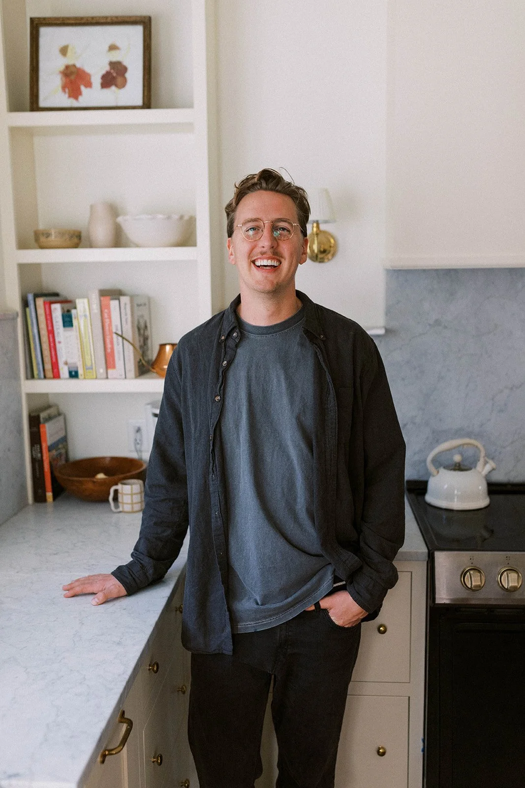 A young man with glasses smiling, standing in a kitchen, leaning on a countertop with shelves filled with books and decorative items behind him, a white kettle on the stove.