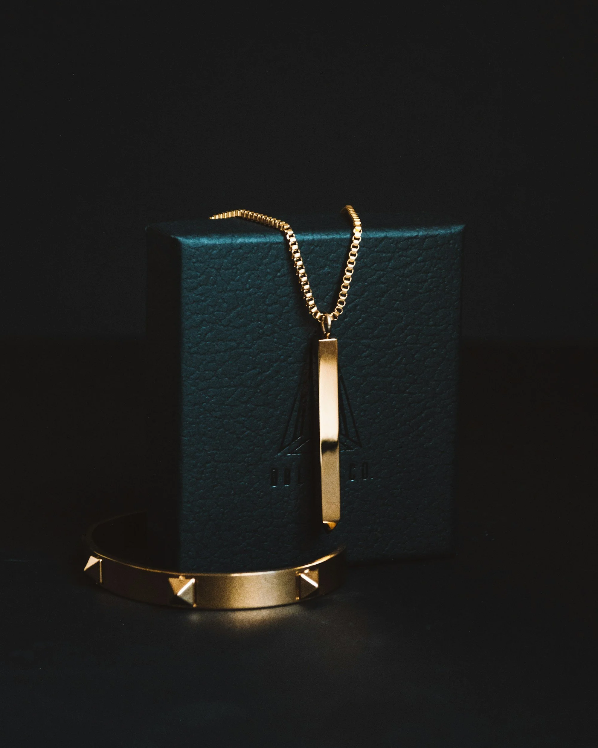 Cadena dorada con colgante rectangular, colocada sobre una caja de color azul oscuro en un fondo negro.