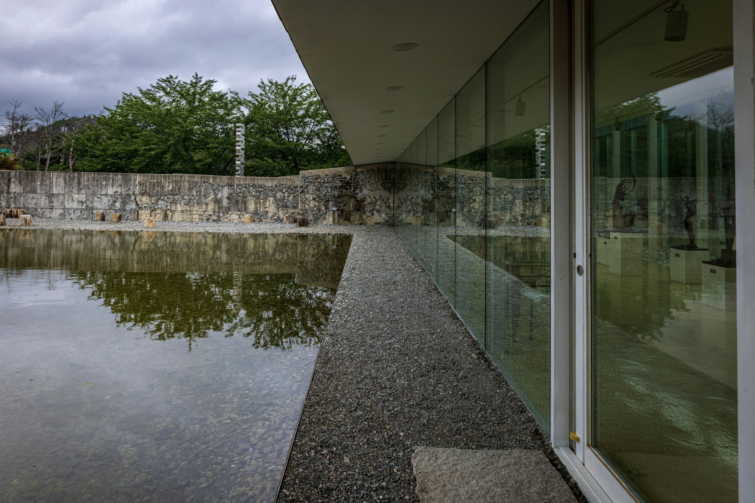Vista lateral de una estructura moderna con paredes de cristal, junto a un estanque de agua y rodeada de árboles y cielo nublado.