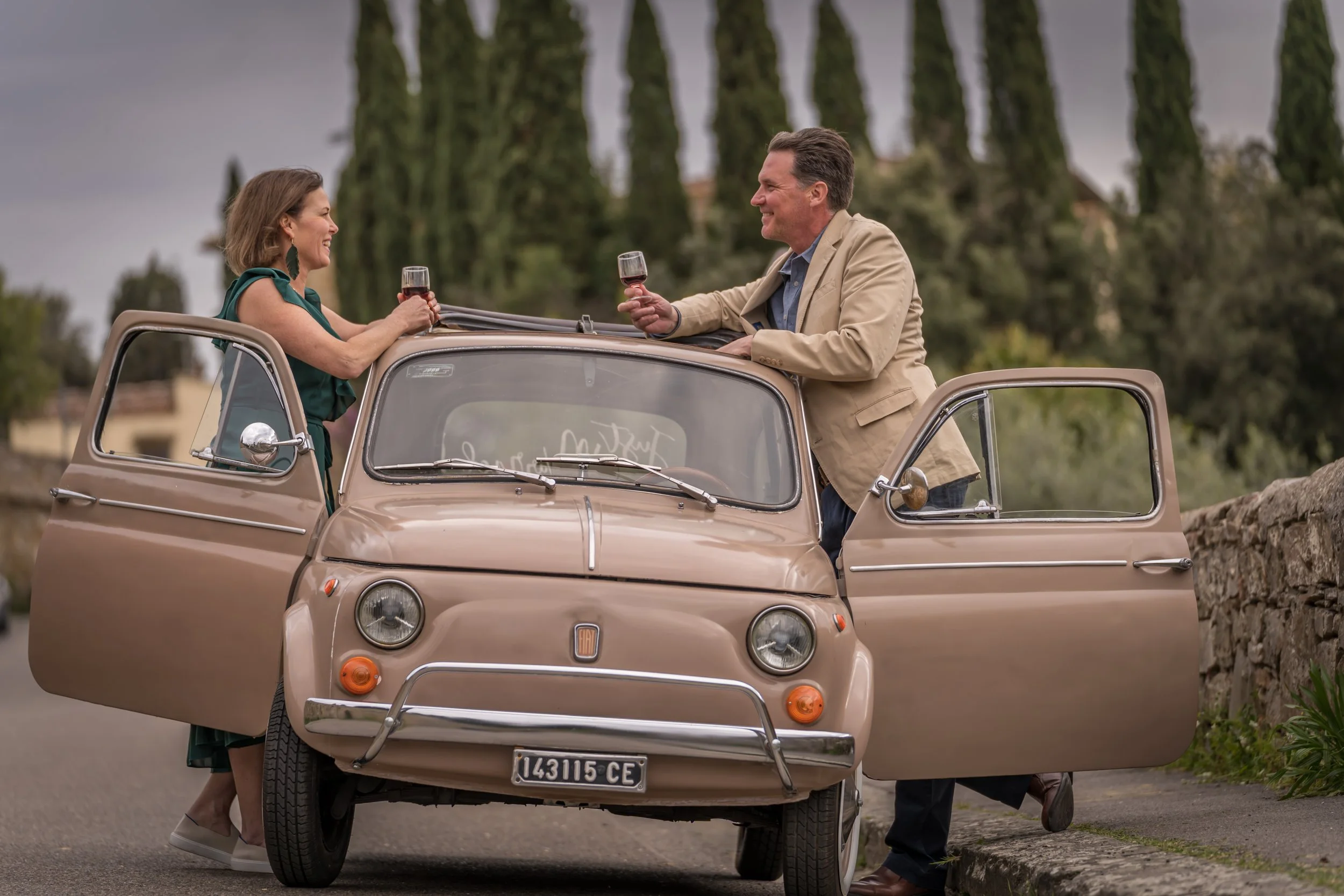 dave and jessica looking at each other over the top of an Italian car, both holding wine glasses.