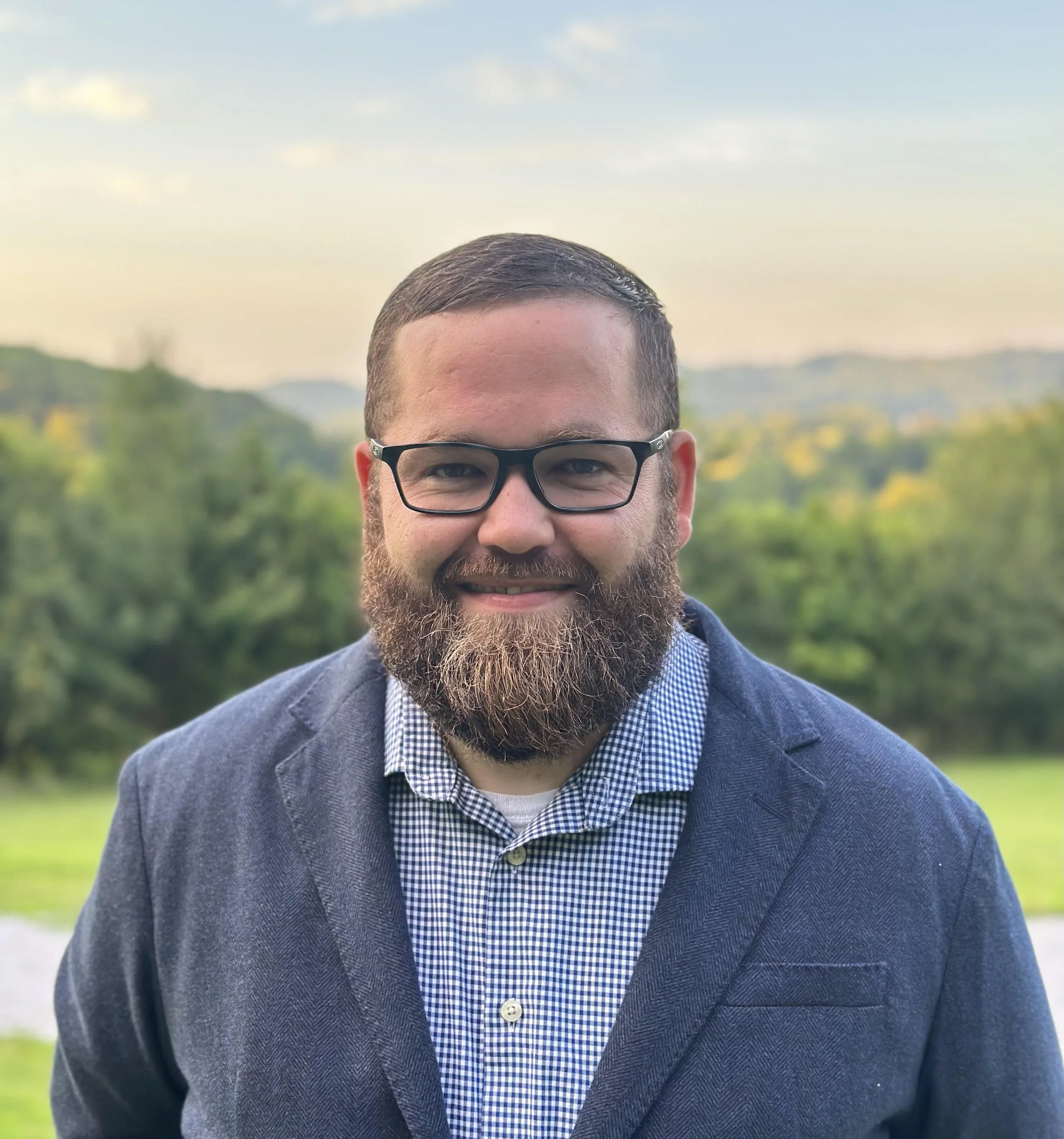Close-up of a smiling man with brown hair, glasses, a beard, wearing a blue and white checked shirt under a dark blazer, outdoors with a blurred green landscape and cloudy sky in the background.