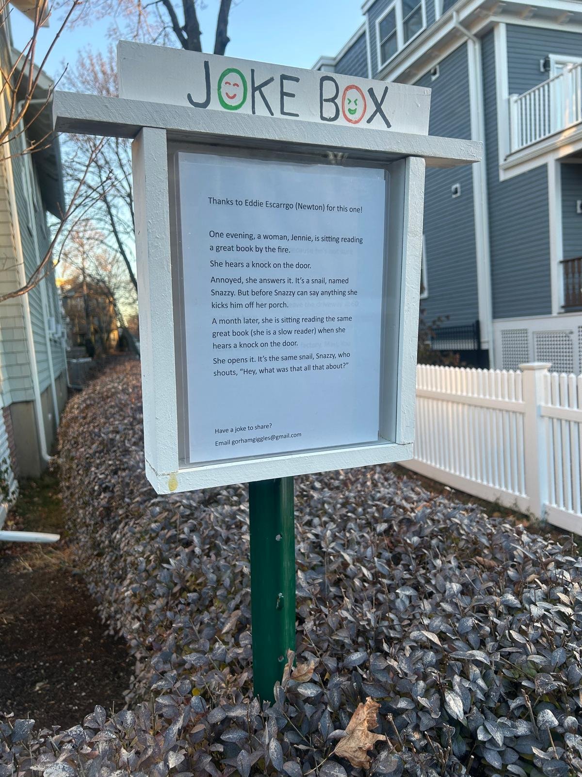 An outdoor white joke box with a sign containing a joke about a snail, sitting on a green post in a residential neighborhood, with houses, trees, and fallen leaves around.
