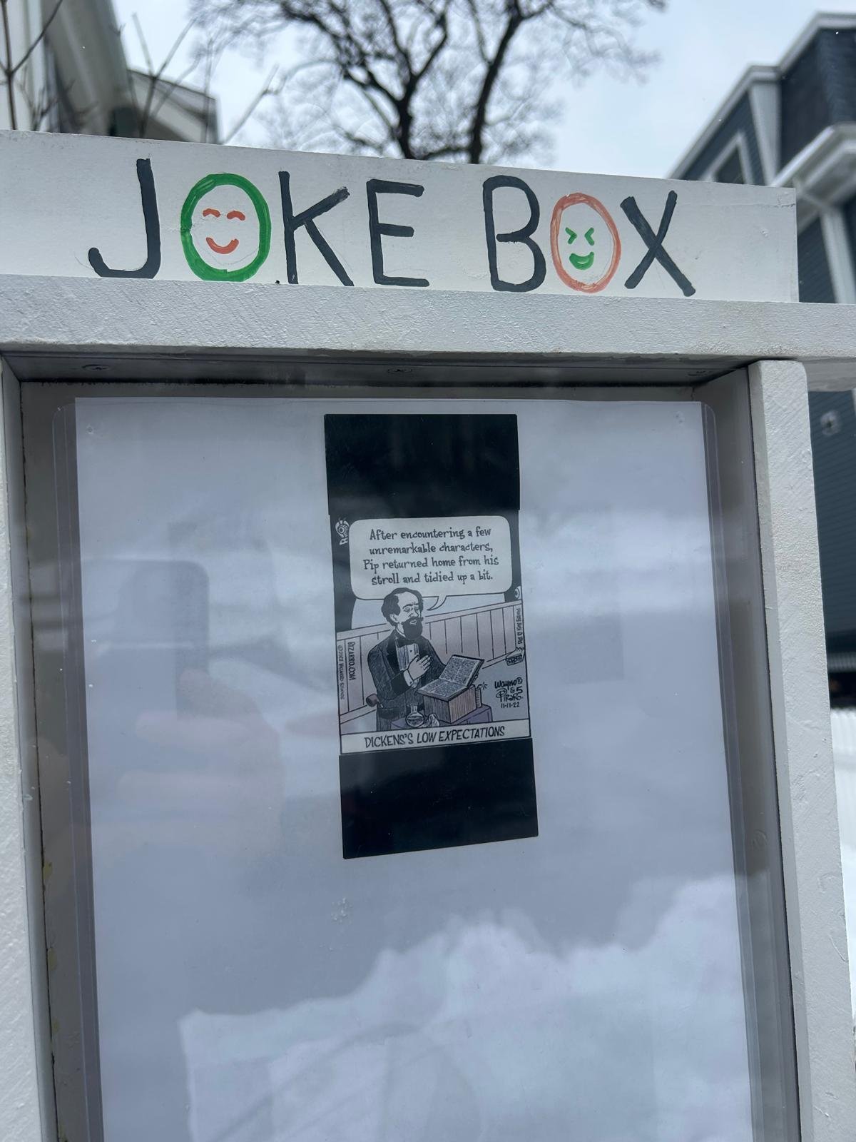 A white box labeled 'Joke Box' with smiley face drawings around the text, positioned outdoors with trees and buildings in the background. Inside the box, there is a black-and-white comic strip with a man reading a book, accompanied by a speech bubble.