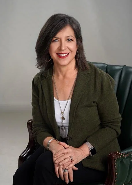 A woman with shoulder-length brown hair, wearing a dark green blazer over a white top, sitting on a green leather chair, smiling at the camera.