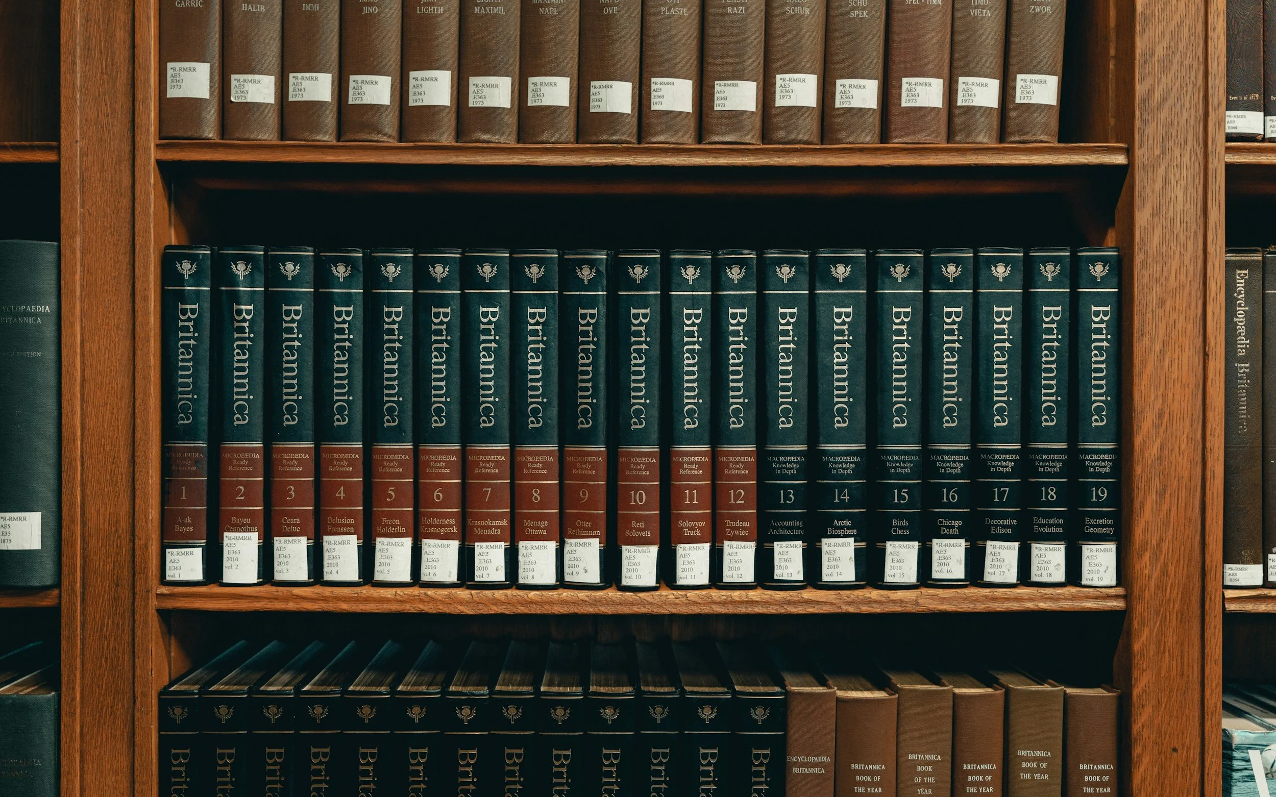 A bookshelf with two rows of books. The top row has brown hardcover books with white labels, and the bottom row has black hardcover books with gold lettering that says 'Britannica.'