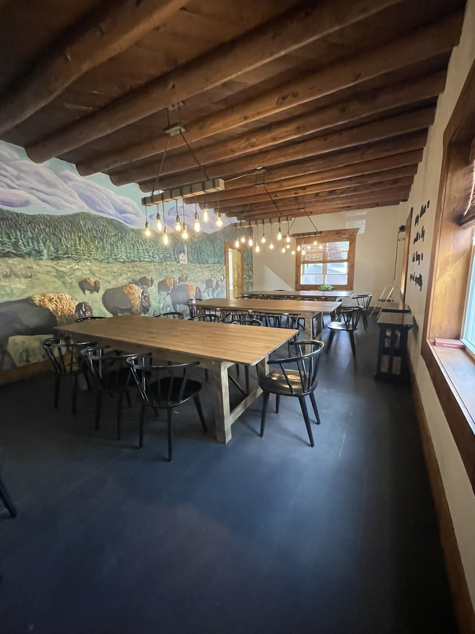 Interior of a rustic restaurant or cafe with wooden ceiling beams, a mural of cows and a landscape on the wall, and hanging Edison-style light bulbs. There are wooden tables and black chairs.