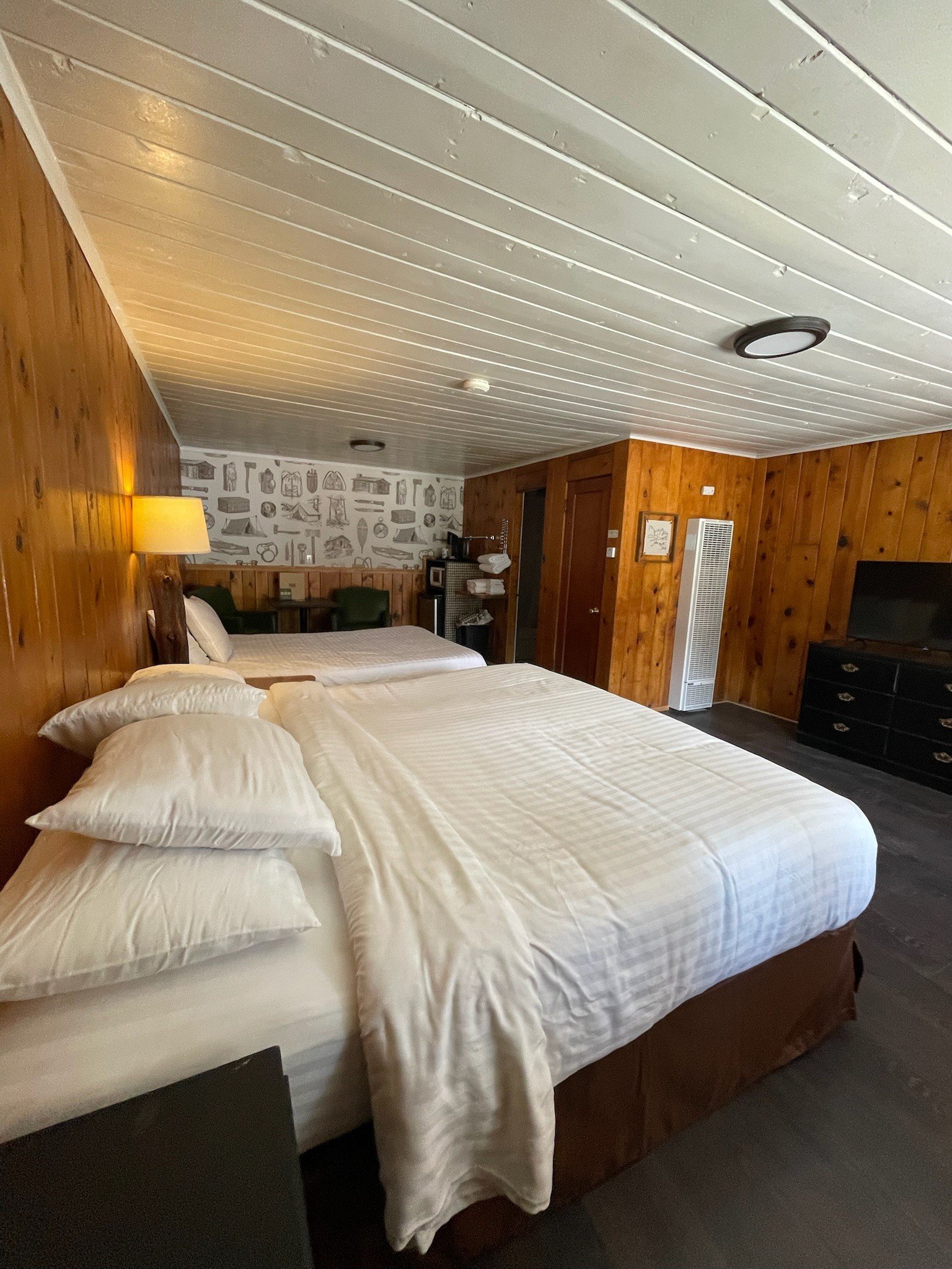 A hotel room with wooden walls and ceiling, two neatly made beds with white bedding, a nightstand with a lamp, a TV, and a patterned wall with various objects.