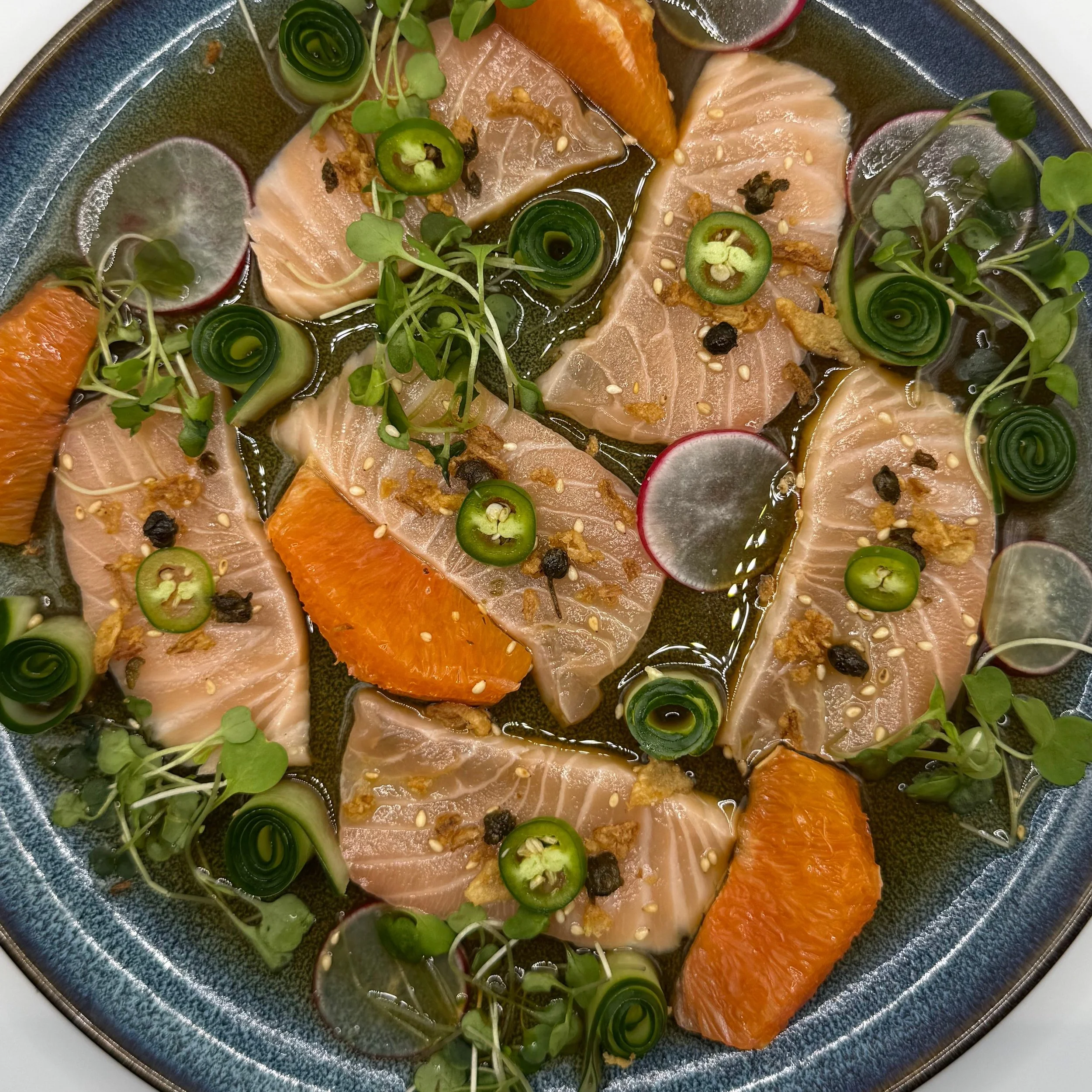 Thinly sliced salmon crudo served with a bright orange-lemon ponzu, layered with citrus segments and crisp cucumber for freshness. Finished with shaved radish, Serrano chile, toasted sesame seeds, and fried capers for salt and crunch, then lifted wit