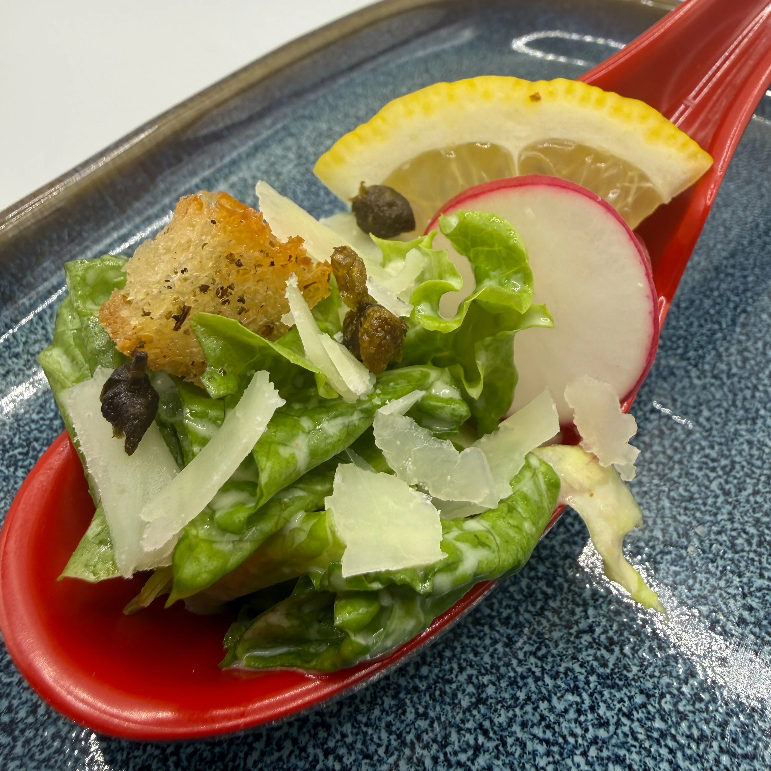 A one-bite tasting of my Signature Caesar salad. Crisp romaine dressed and nestled in a tasting spoon with a petite golden crouton, sharp Parmesan flake, and crispy capers.
