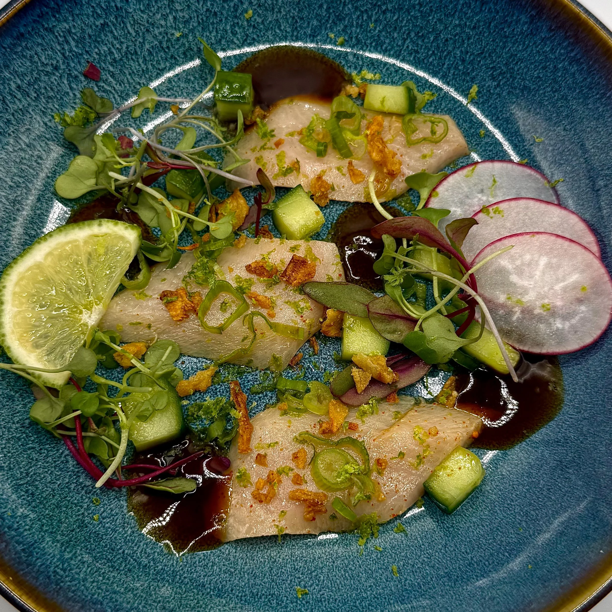 Yellowtail Crudo topped with crisp cucumber, fresh micro-greens, and a savory toasted garlic crunch. Finished with a splash of spicy ponzu, lime zest, and radish for a bright, acid-forward finish.