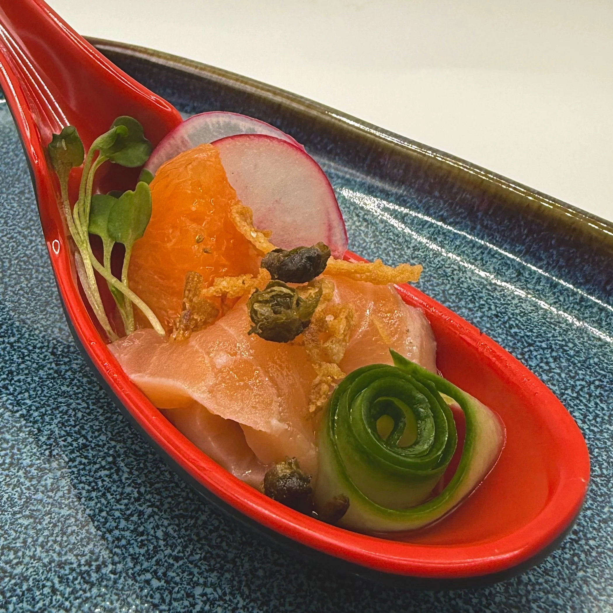 A vibrant tasting spoon featuring hand-pressed salmon nigiri and a hand-torn orange segment, topped with crispy aromatics and a precision cucumber ribbon. 