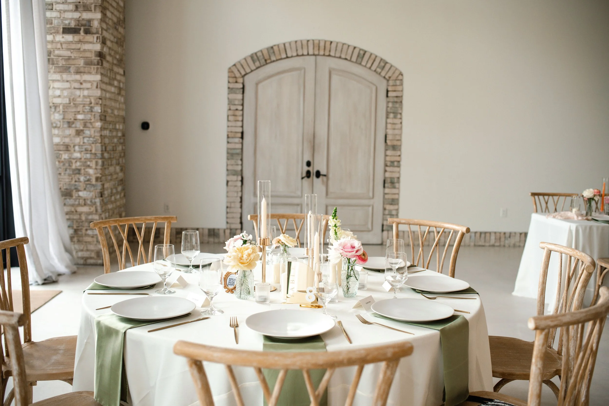 A round table set for a formal event with white tablecloths, floral centerpieces, candles, wine glasses, and place settings, surrounded by wooden chairs, in a room with a brick wall and double doors.