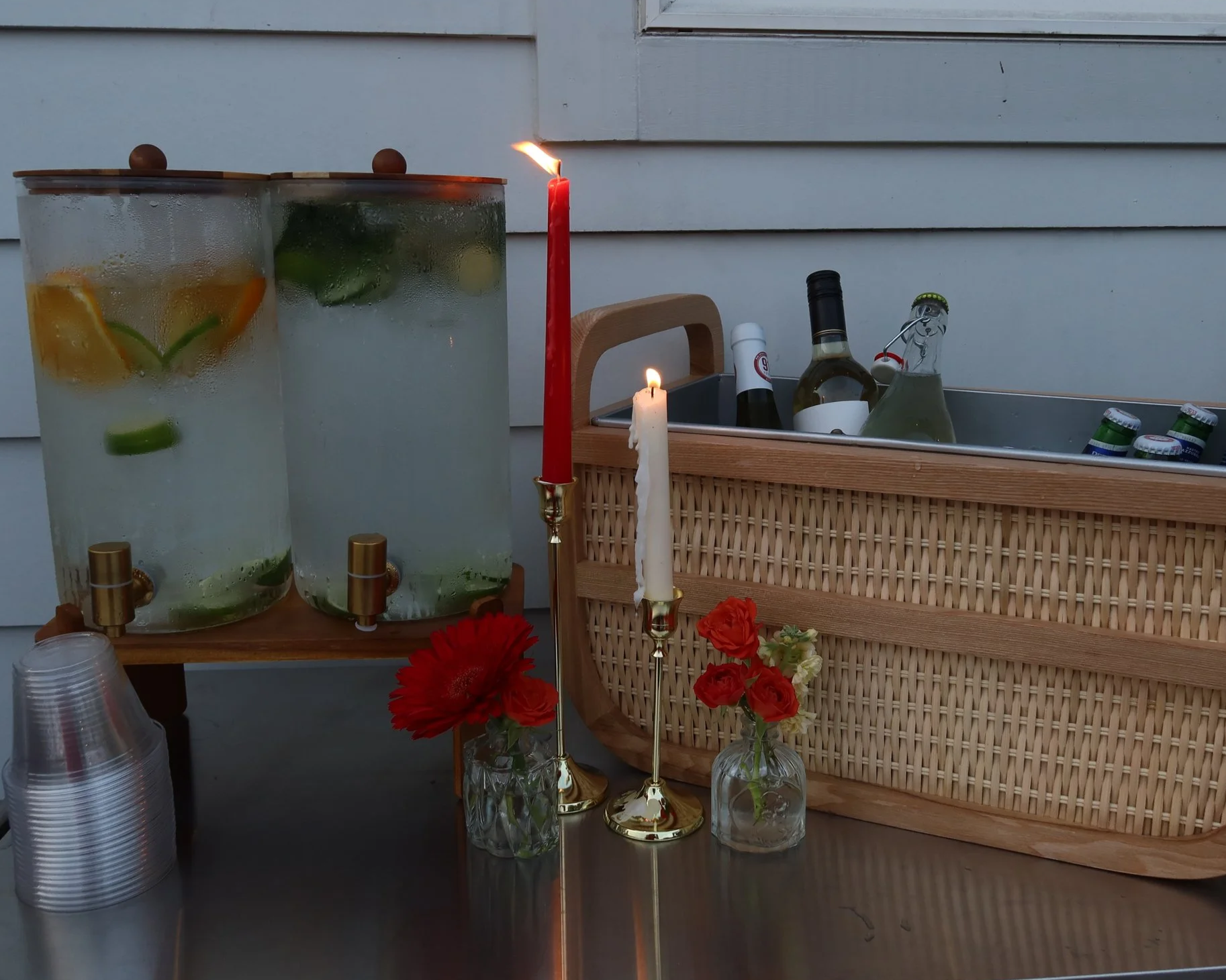 Two glass beverage dispensers with fruit-infused water on a wooden stand, candles, a basket, bottles, and flowers on a table near a light gray wall.