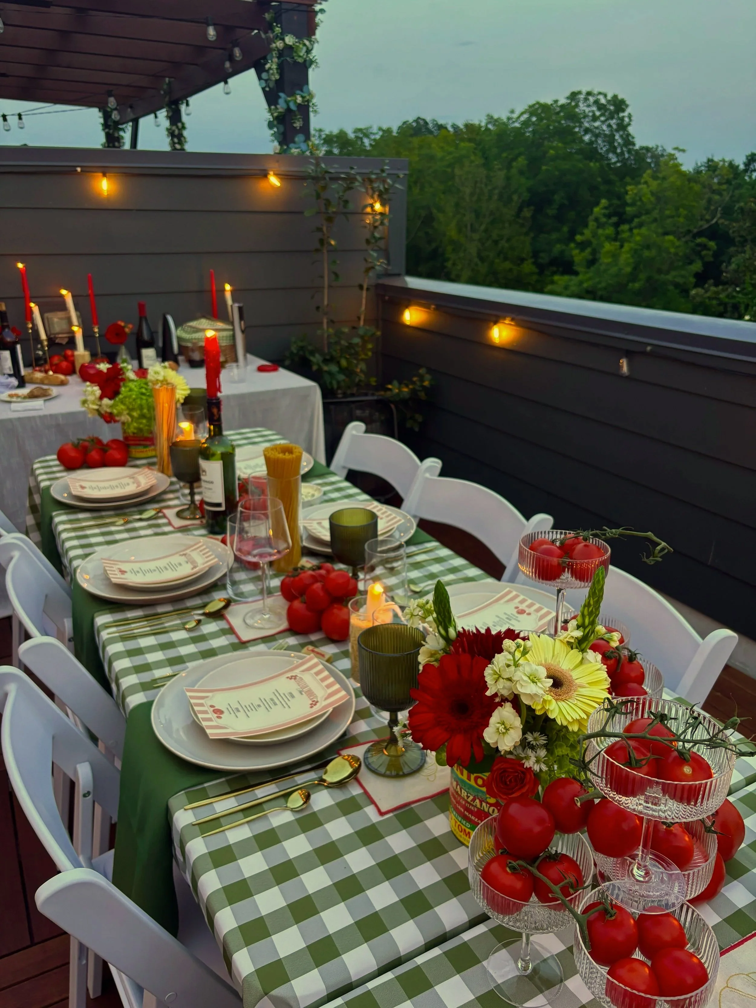A decorated outdoor dining table with green and white checkered tablecloth, floral centerpieces, wine bottles, candles, and tomatoes in glass bowls, set on a balcony with string lights and a view of trees in the background.