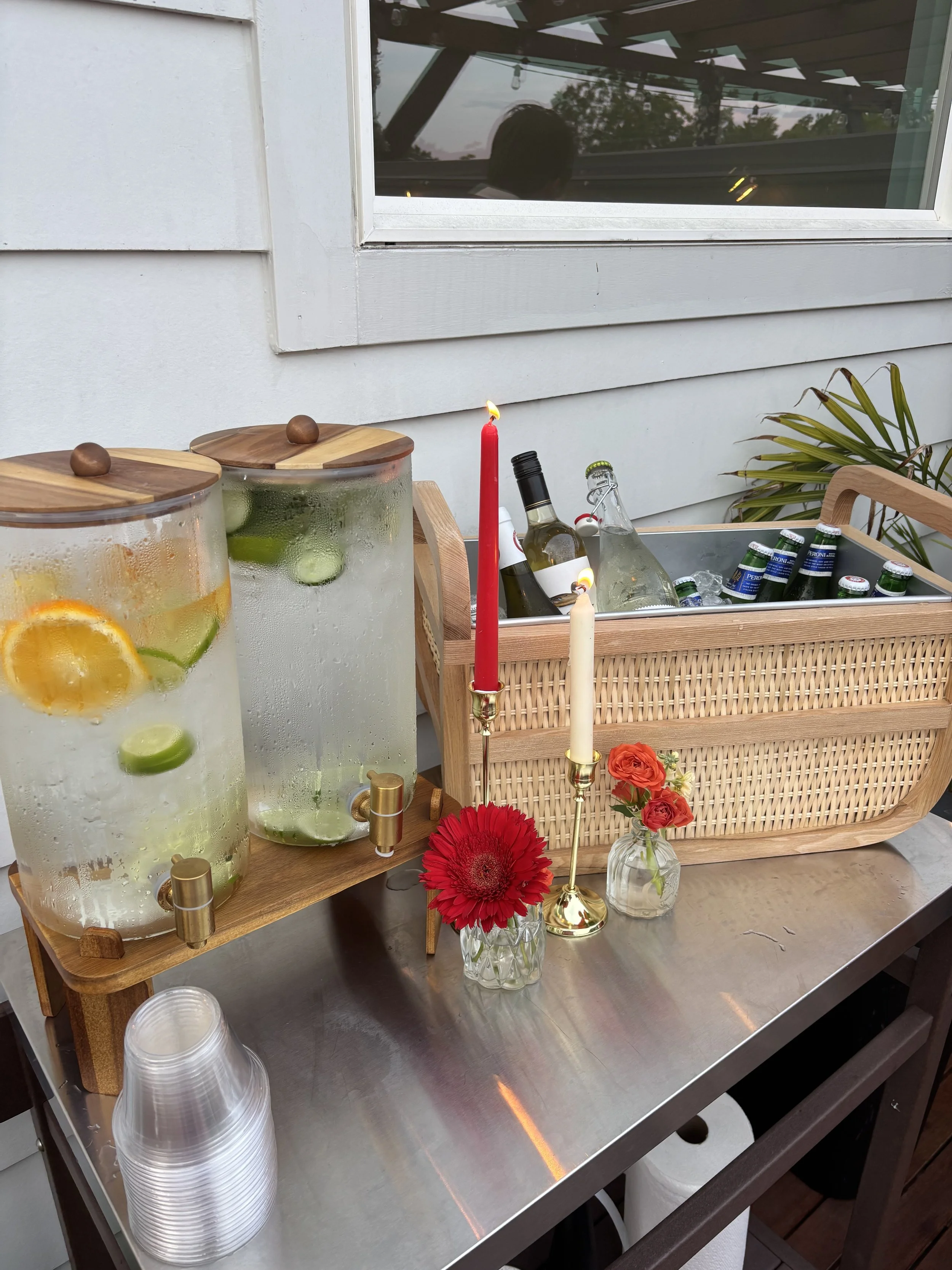 A table setup with two large glass pitchers filled with lemon and lime infused water, a red candle, white and red candles, and a small bouquet of flowers in a glass vase. There is also a cooler filled with bottles of sparkling water or soda, an arran