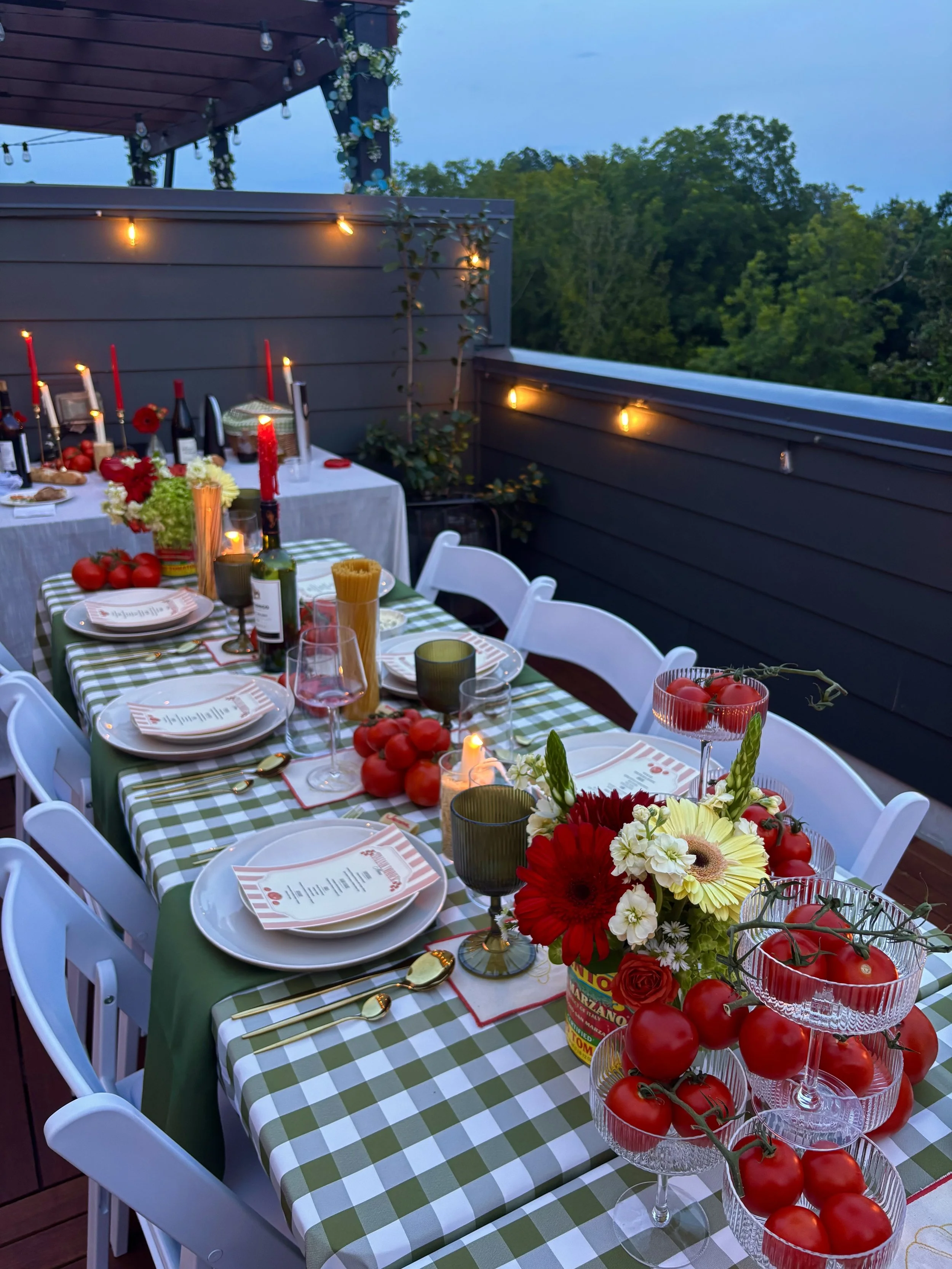 An outdoor dining table with a green and white checkered tablecloth, decorated with flowers, candles, wine bottles, wine glasses, plates, and tomatoes, set for a meal on a balcony or patio with string lights and surrounded by trees.