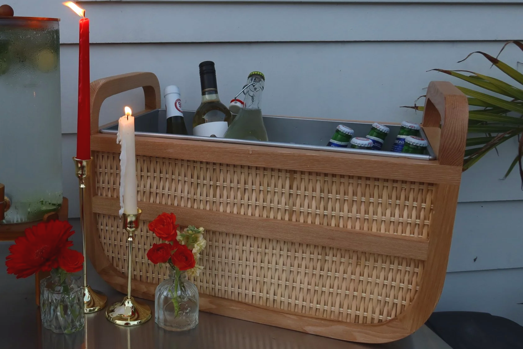 A wicker cooler filled with bottles of wine, a glass bottle of soda, and cans of beer. There are two lit candles on either side and a flower arrangement with red and white flowers in front.