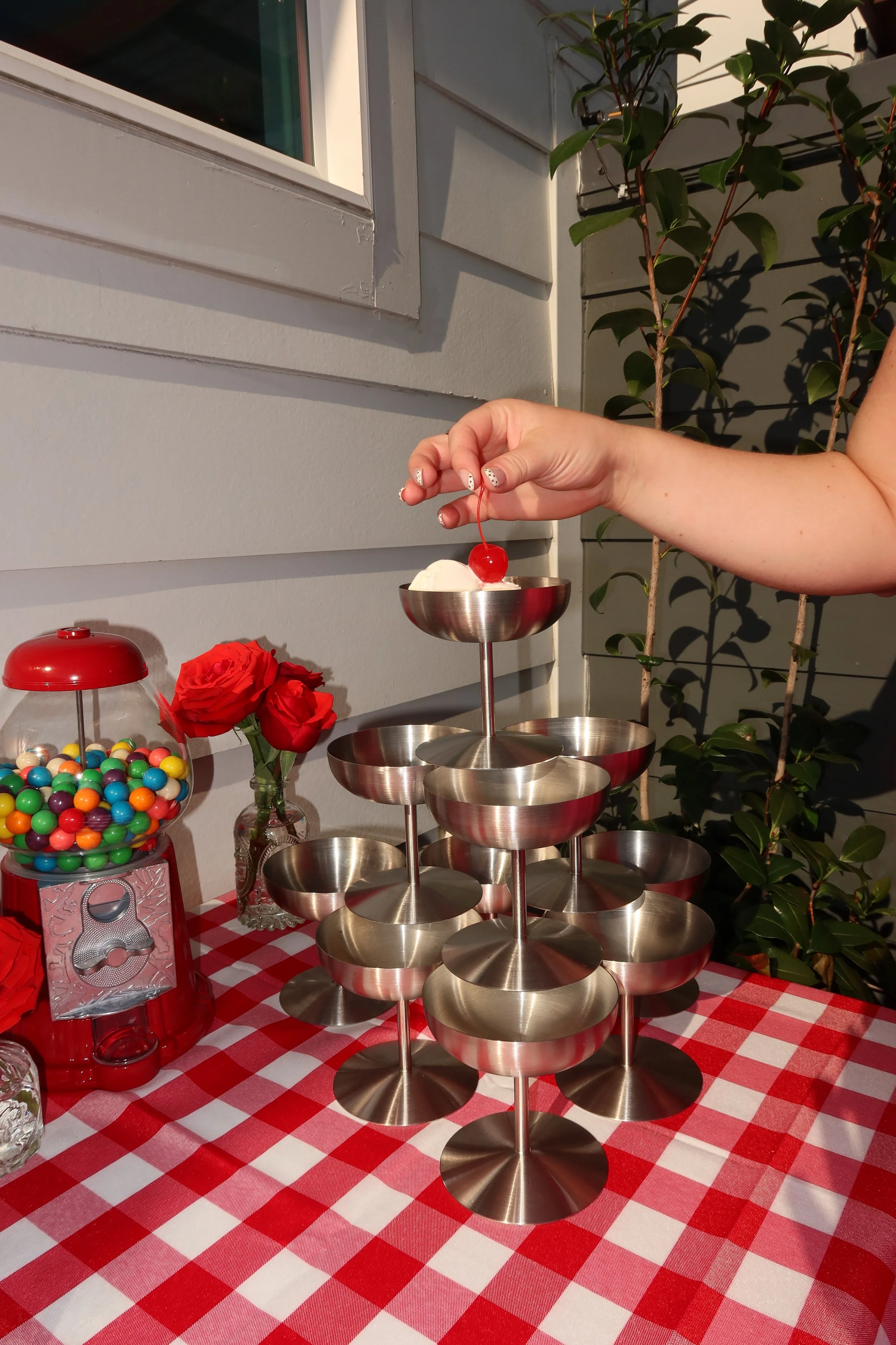 A person is holding a maraschino cherry over a tiered stainless steel dessert stand. The setting is a table with a red and white checkered tablecloth, decorated with a jar of colorful candies, red roses, and an ice cream scoop.