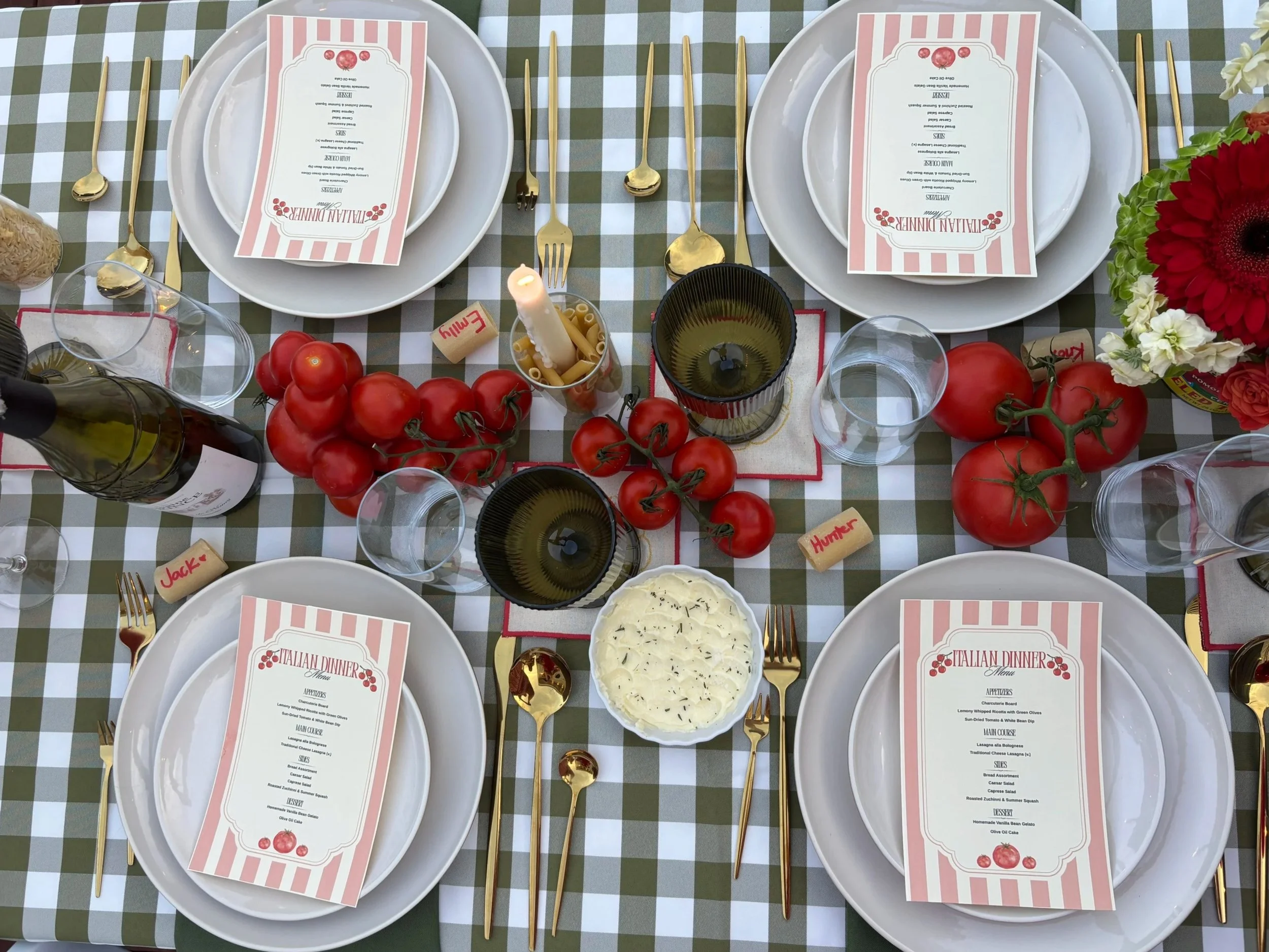 Table setting for an Italian dinner with white plates, gold utensils, wine glasses, and a menu card on a green and white checkered tablecloth. The table is decorated with tomatoes and flowers.