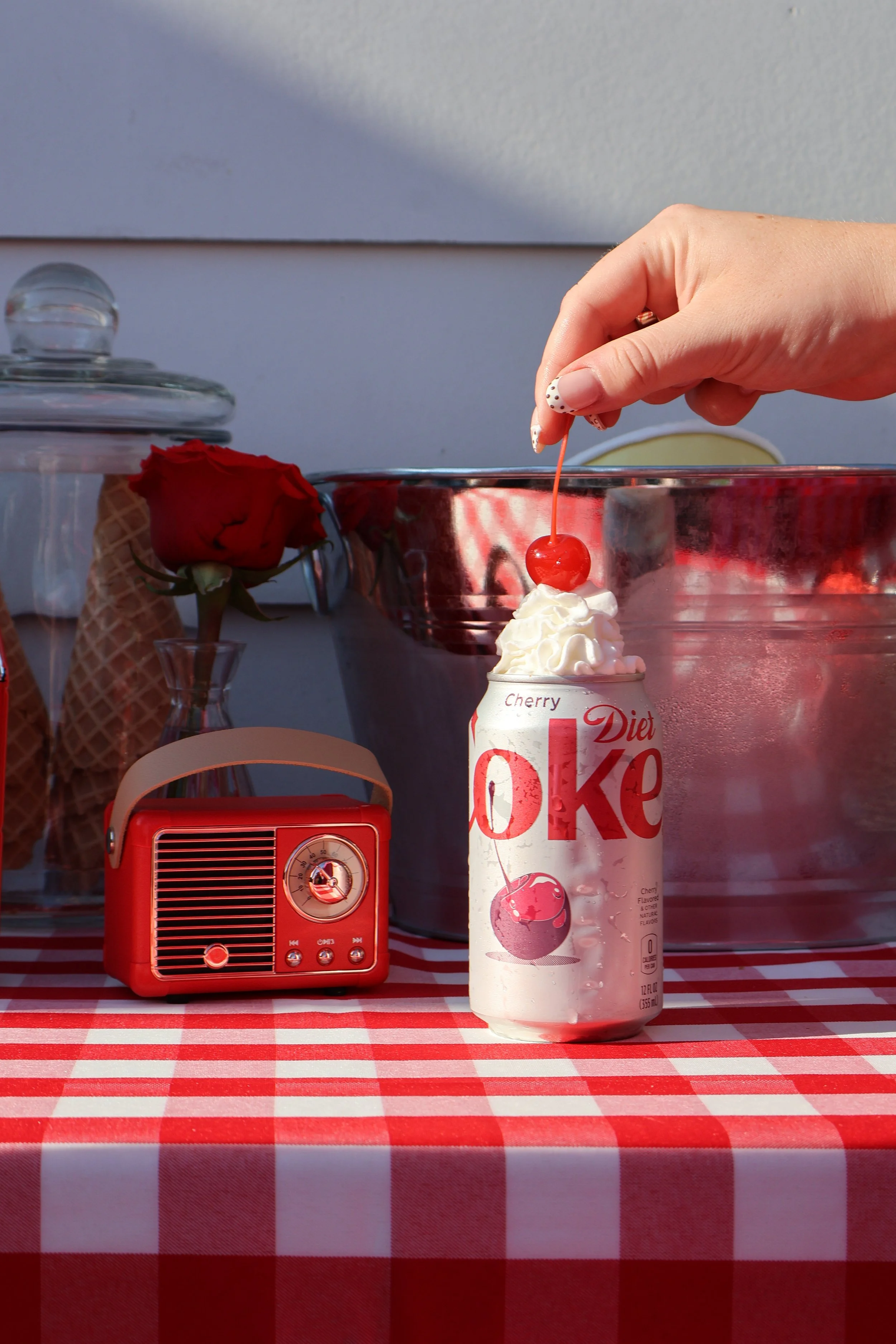 A can of Diet Coke Cherry flavor with whipped cream and a maraschino cherry on top, a small red radio, a red rose in a glass, ice cream cones, and a tray of ice in the background on a red and white checkered tablecloth.