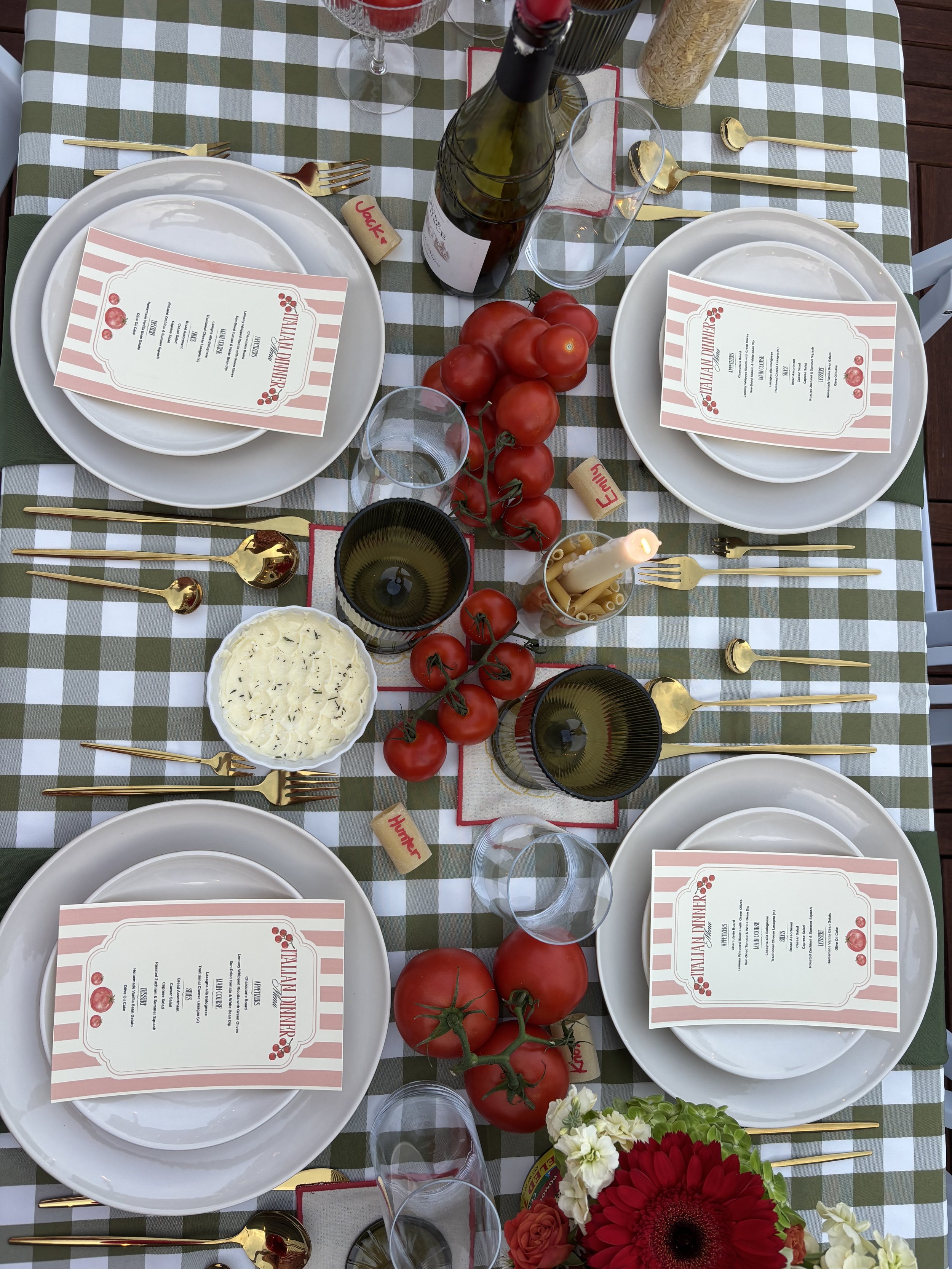 A decorated table set for a meal with white plates, gold utensils, wine glasses, a bottle of wine, and a menu on top of each plate. There are tomatoes, flowers, a candle, and various food items such as pasta and cheese on the table. The tablecloth ha