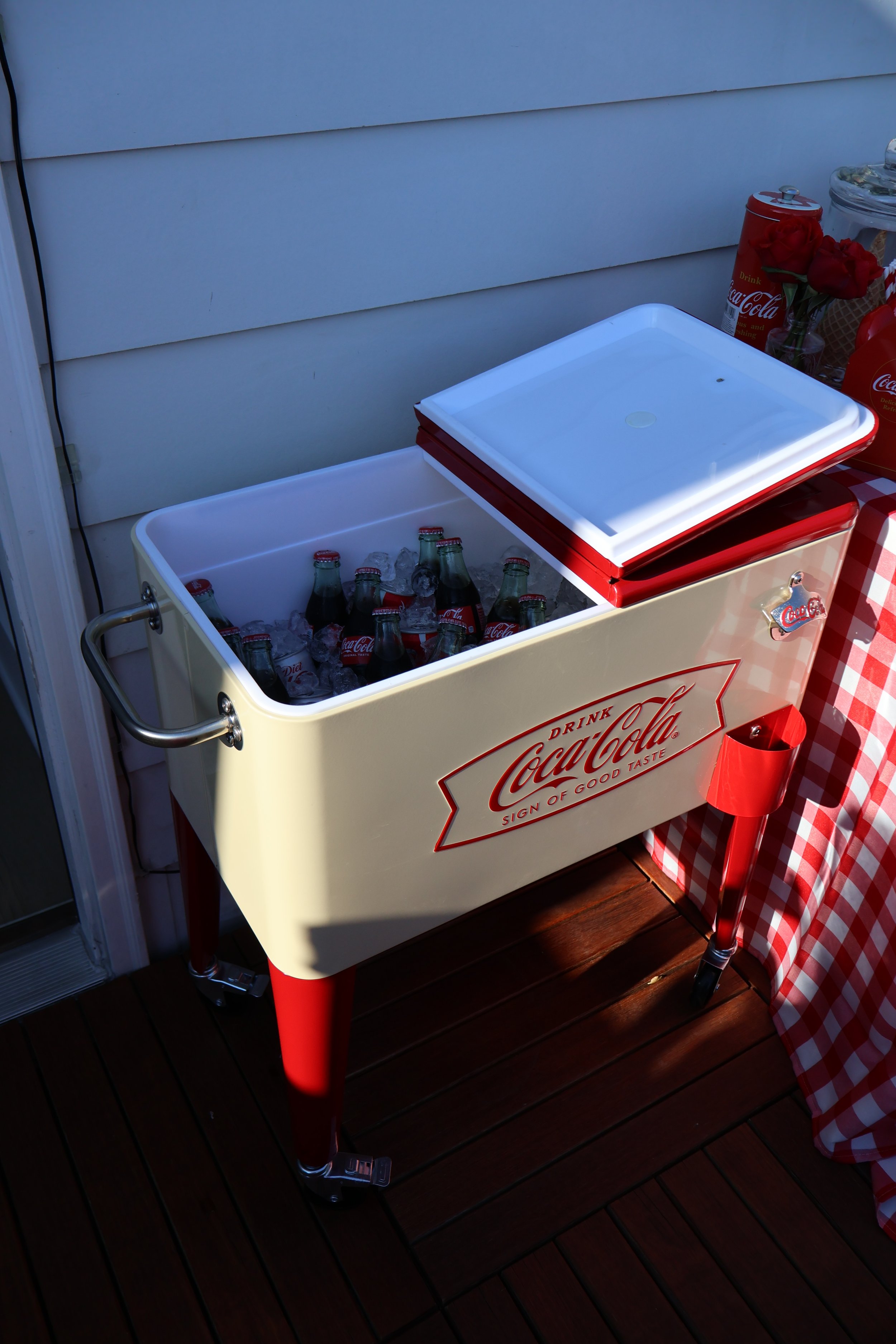 A vintage Coca-Cola cooler filled with ice and glass Coca-Cola bottles. The cooler is cream-colored with red accents and sits on a wooden deck beside a red-and-white checkered tablecloth.