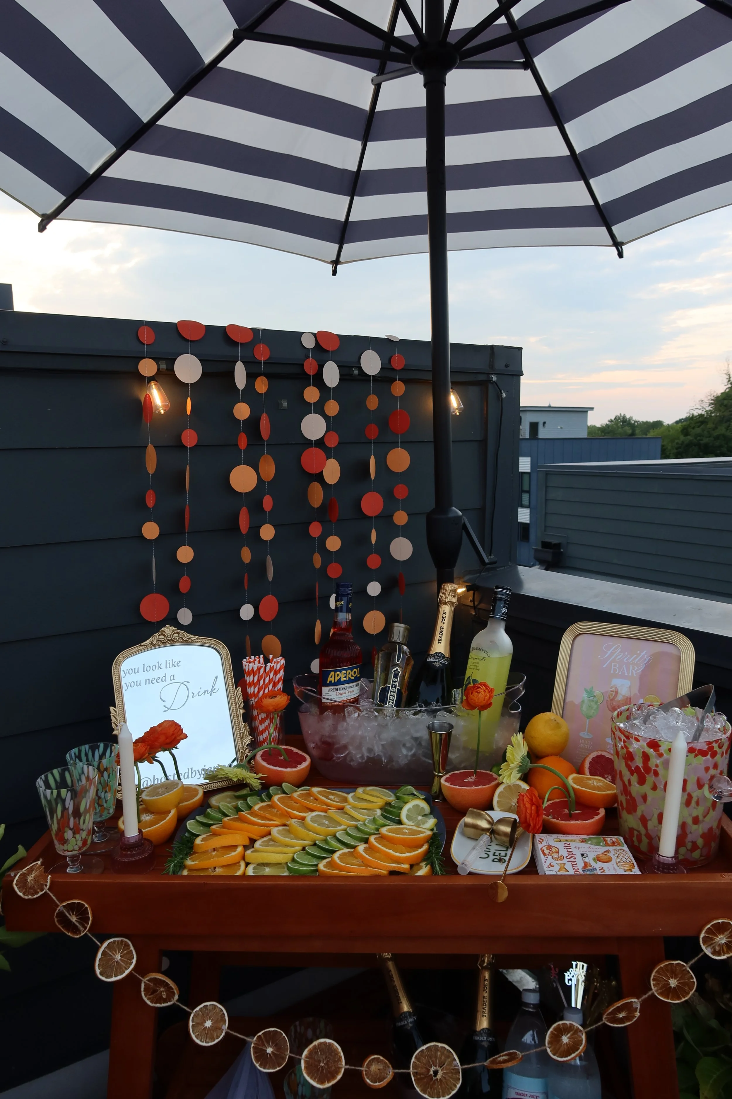 Outdoor bar counter decorated with a striped umbrella, orange garland, various bottles of alcohol, a bowl of ice with lime, lemon, and grapefruit slices, and small framed signs.