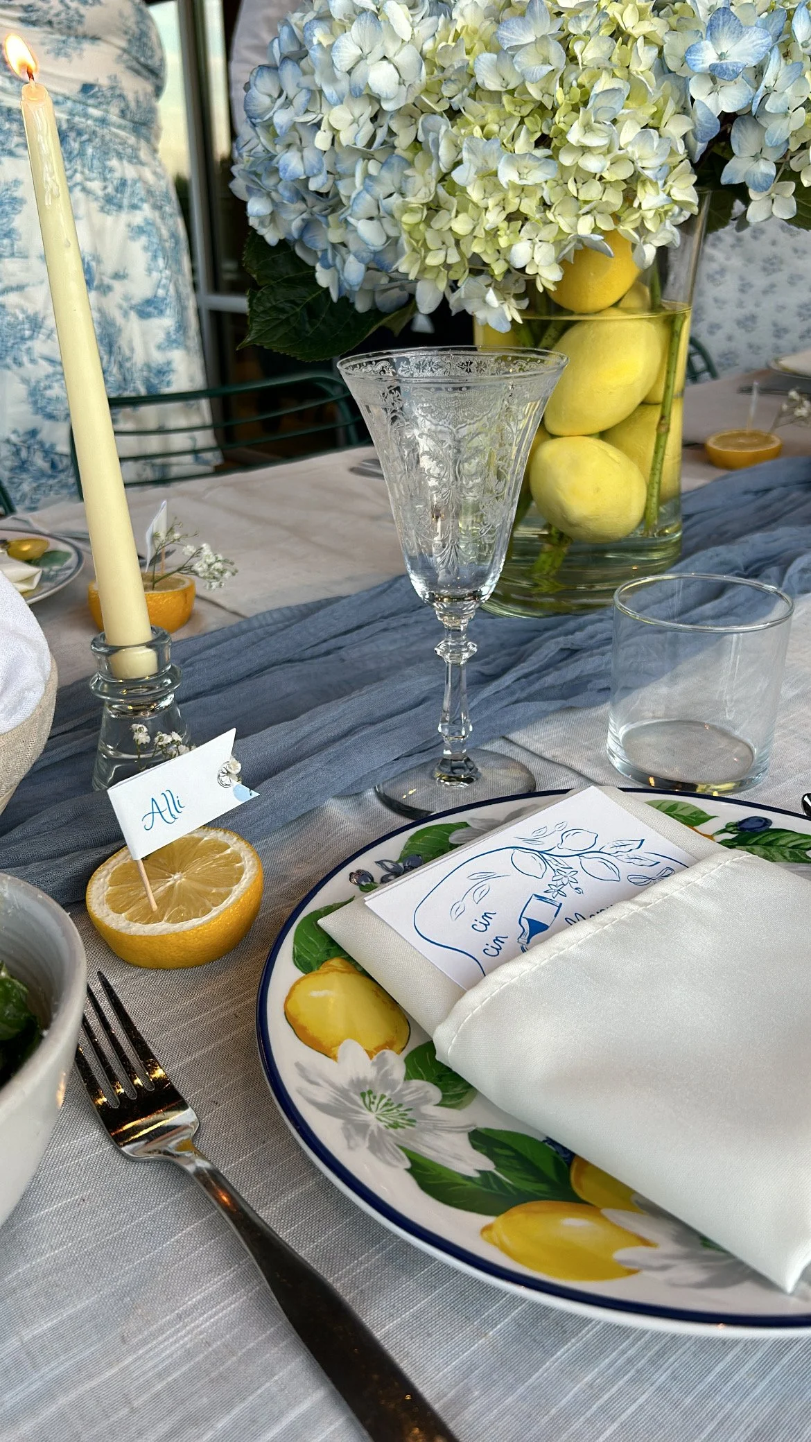 A table set for a meal with a white tablecloth, floral-patterned plate, white napkin, glassware, and a small lemon half with a name tag that says 'Ali'. There is a tall candle, a large floral arrangement with white and blue hydrangeas, and a bowl of 