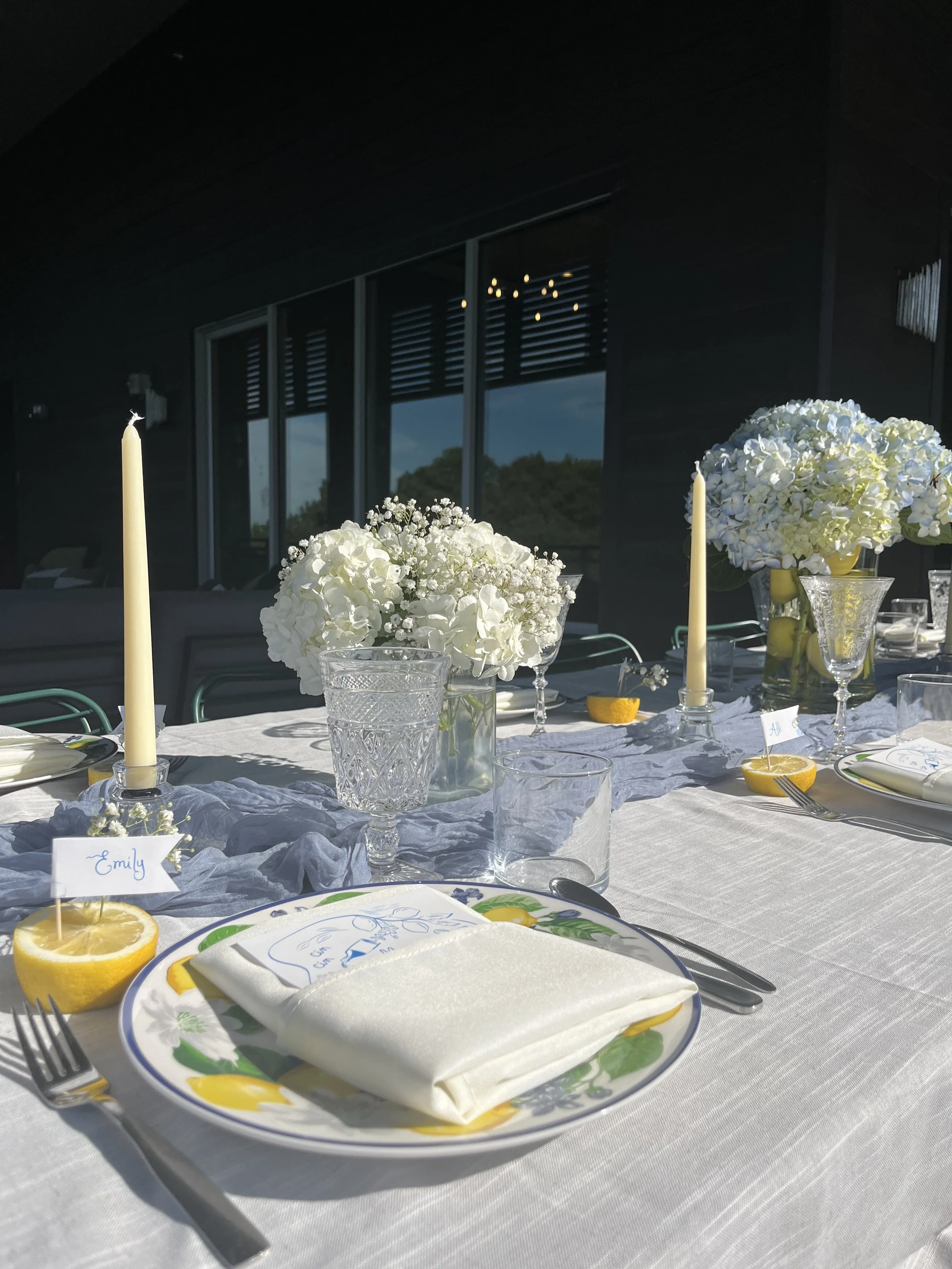A decorated table set for a meal with floral centerpieces, candles, lemon slices, and place settings with plates, napkins, and utensils, in a room with large windows.