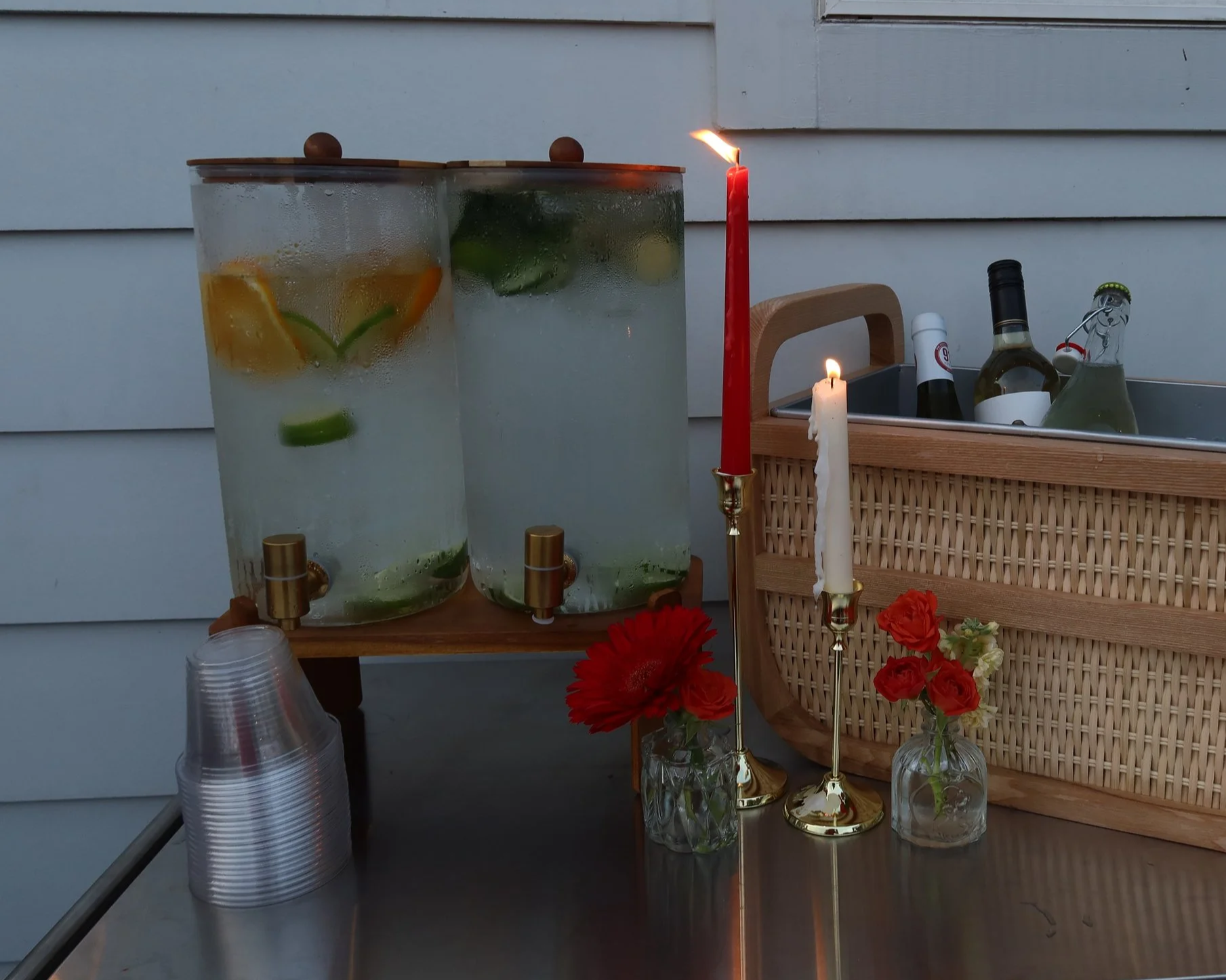 Two large glass drink dispensers filled with citrus slices and mint, placed on a wooden stand. There are lit candles, a bottle of wine, and flowers in small vases on a table. A basket with bottles and a white wall are in the background.