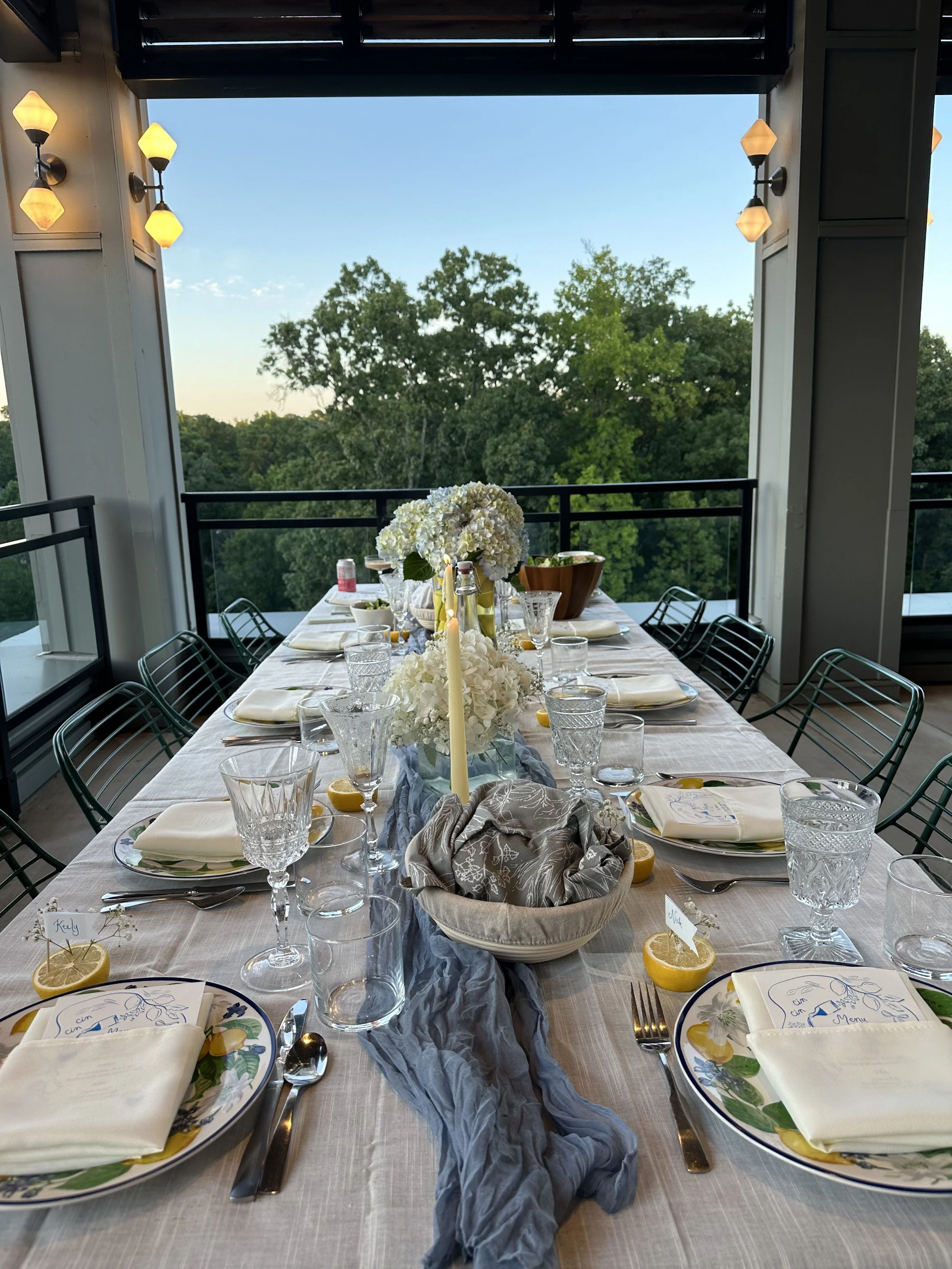 A decorated dining table set indoors with a centerpiece of white hydrangeas, surrounded by glassware, plates, napkins, and lemon slices, with outdoor trees visible through large windows in the background.