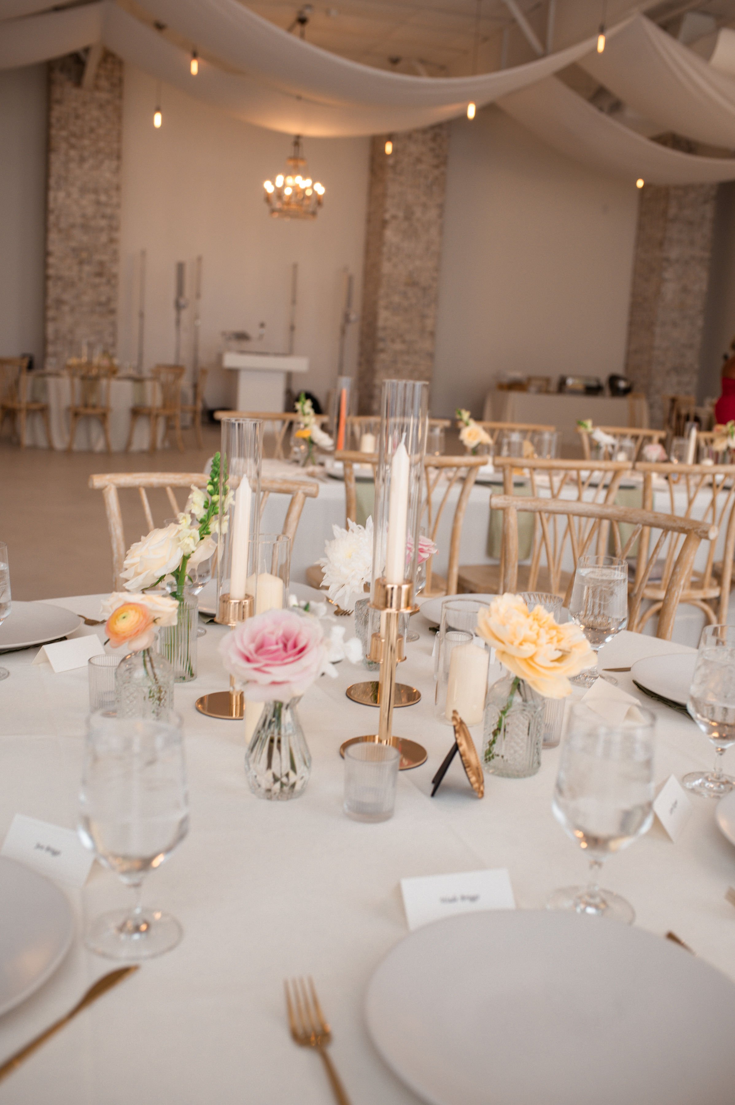 Wedding reception table decorated with pink and white flowers, candles, and glassware in a rustic venue with exposed brick walls, wooden chairs, and hanging string lights.