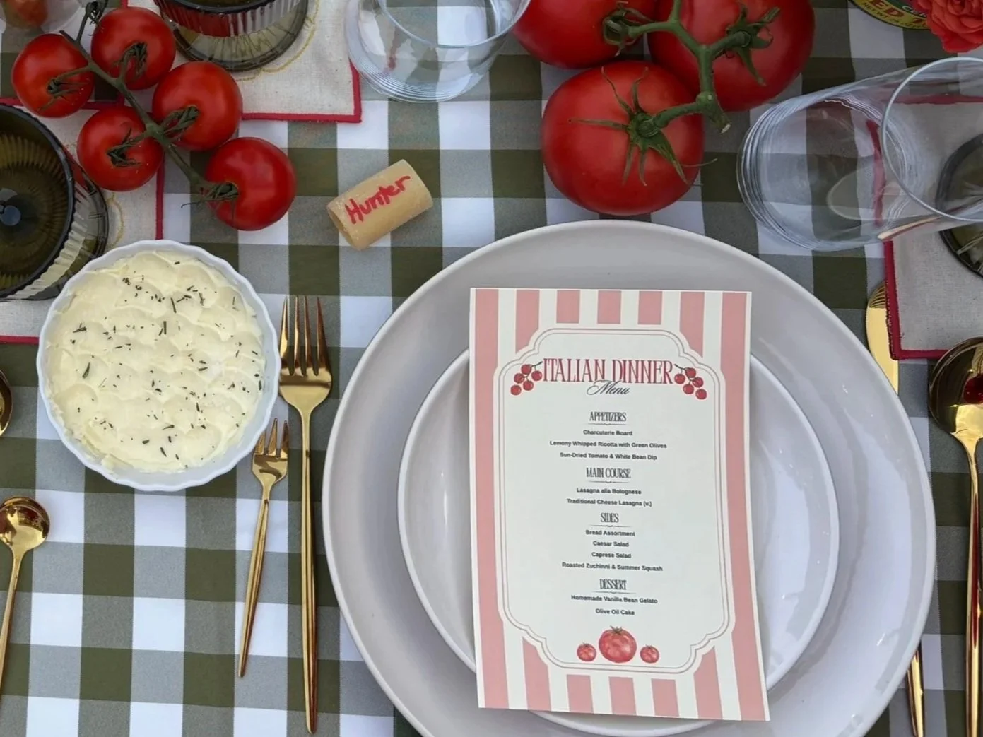 A table set for an Italian dinner with a checkered tablecloth, tomatoes, a menu, mashed potatoes, and gold utensils.