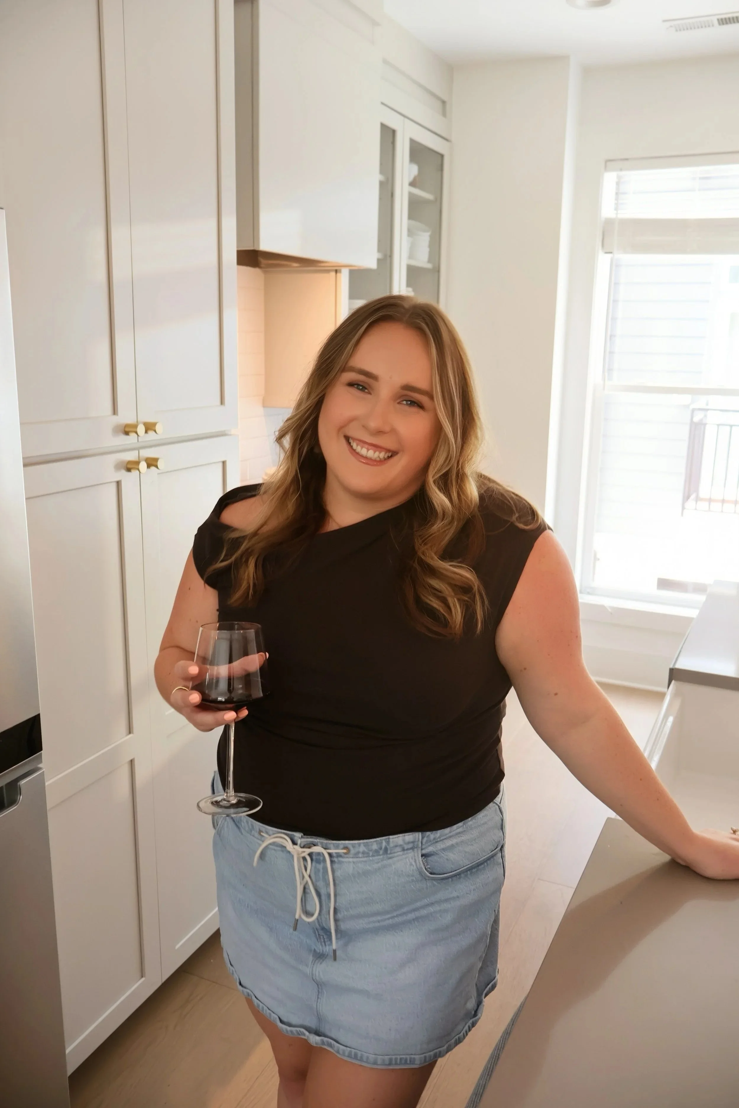 A woman smiling, holding a glass of red wine, standing in a bright kitchen with white cabinets and large windows.