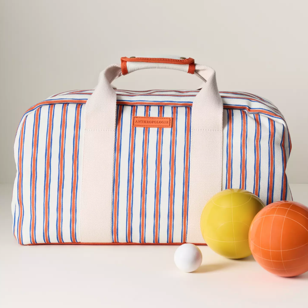 A striped travel bag with the brand name 'Anthropologie' on a leather patch, placed alongside three small balls in yellow, orange, and white, on a white surface against a plain white background.