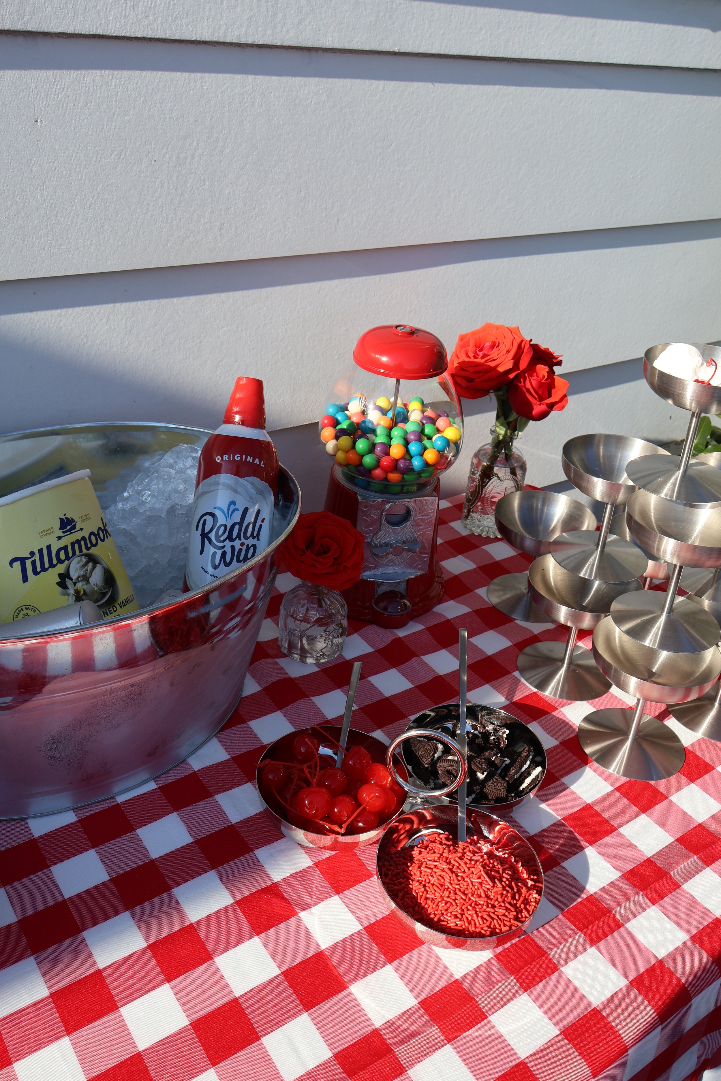 Table with red and white checkered tablecloth holding various desserts including cherries, chocolate pieces, and sprinkles, as well as a bowl of colorful gumballs, a bouquet of red roses, and a stainless steel three-tiered serving stand, outside agai