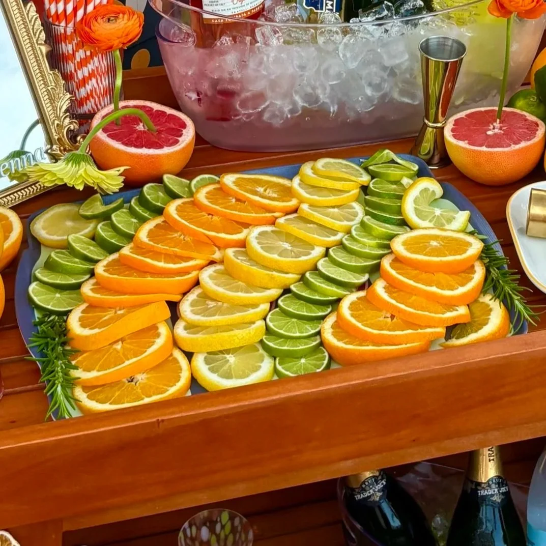 A tray of sliced citrus fruits including oranges, lemons, and limes arranged in rows. Two halves of grapefruit with flowers on top are positioned behind the tray. There are bottles of sparkling wine and a bucket of ice in the background, along with a decorative mirror, flowers, and other party items.