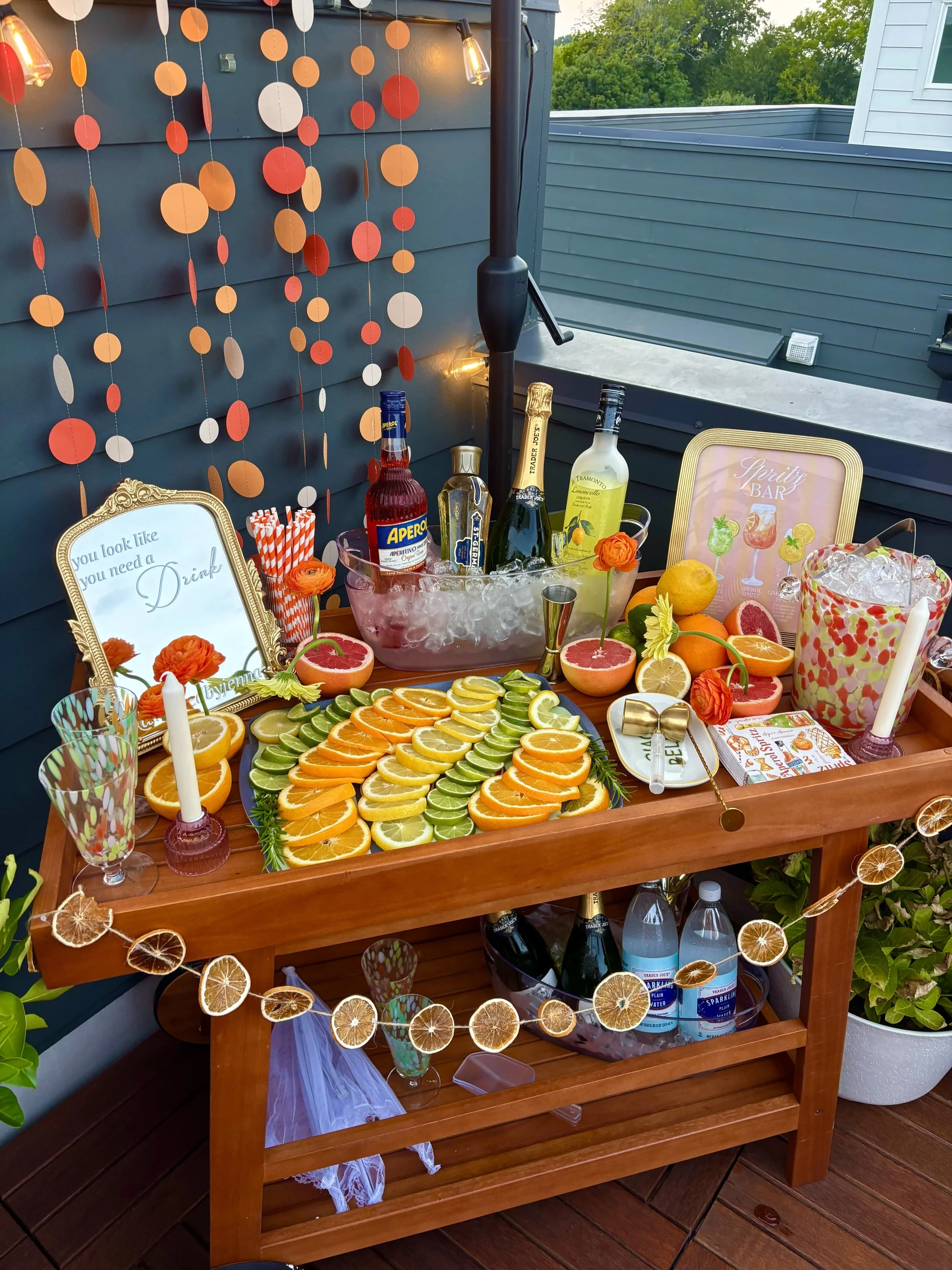A bar setup on a wooden table with sliced citrus fruits, bottles of alcohol, a framed sign, flowers, and a garland of dried citrus slices.