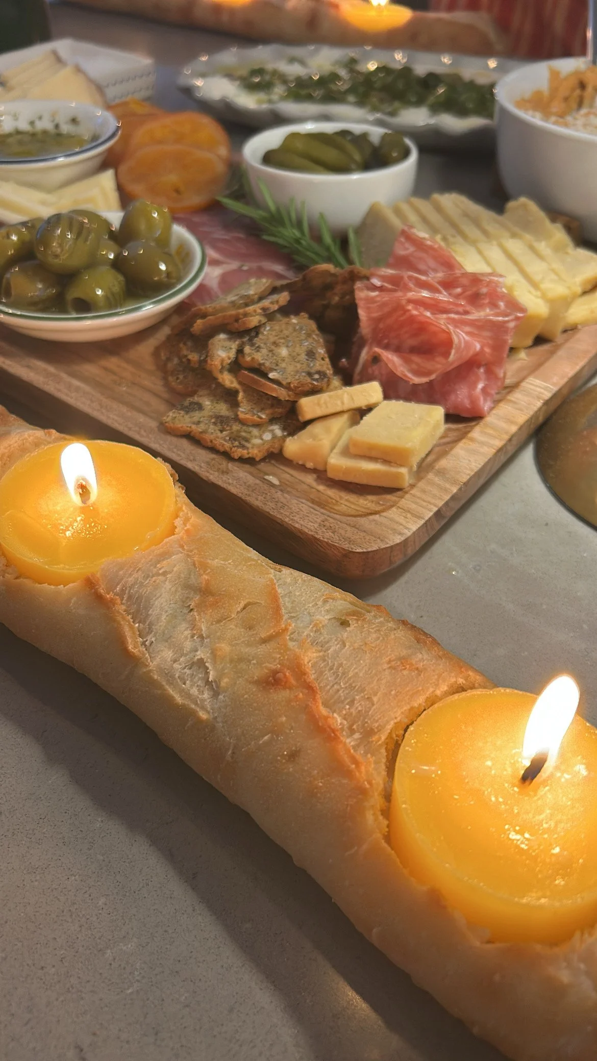 A charcuterie board with various cheeses, cured meats, pickles, and bread. There is a rolled baguette with candle flames on top.