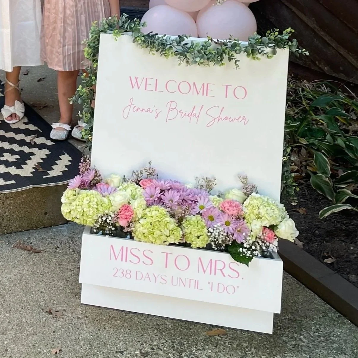 A decorative sign welcoming guests to Jenna's bridal shower, with floral arrangements in pastel colors at the base. The sign indicates 238 days until the wedding.