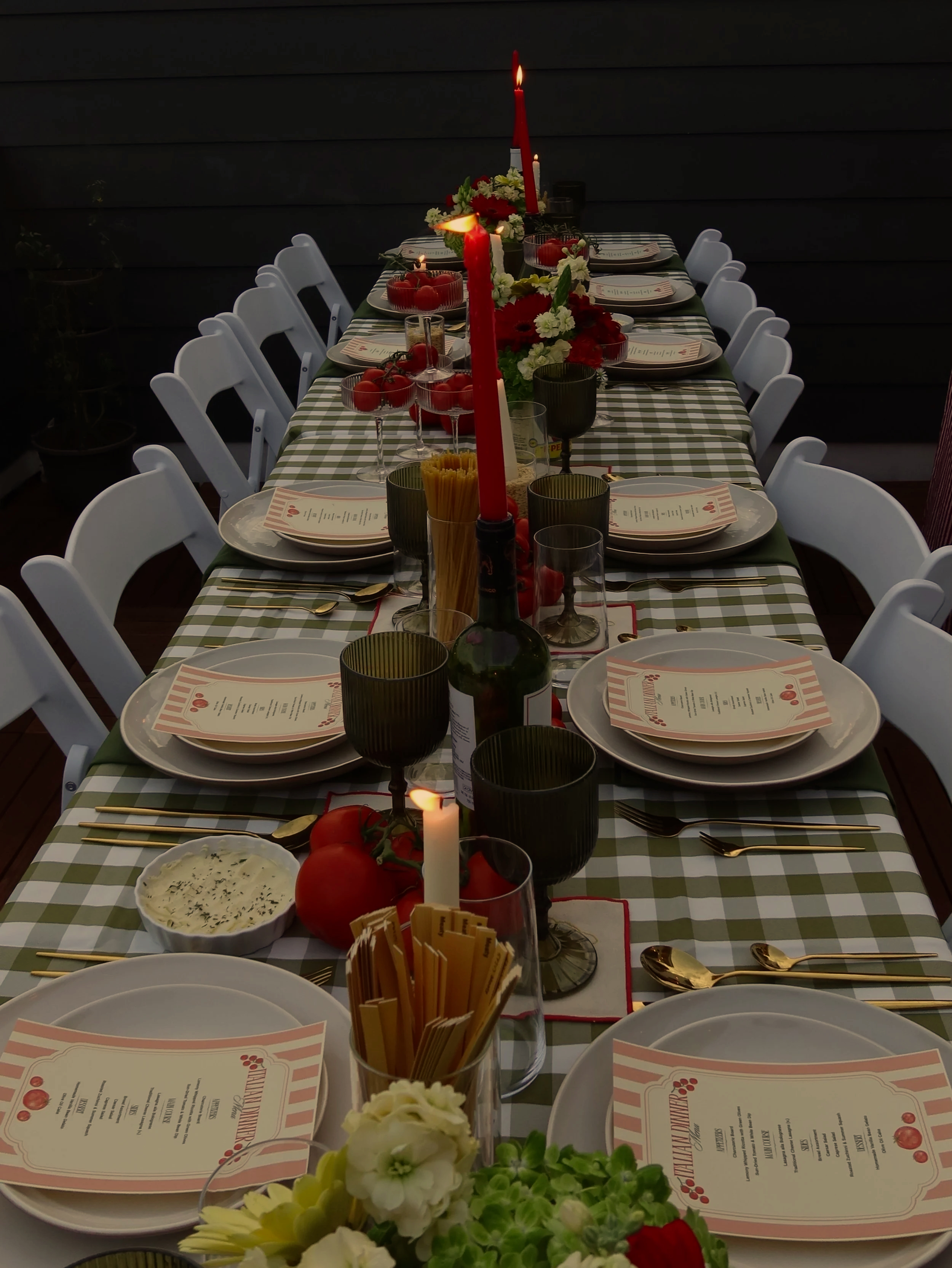 A long dining table set for a meal with white chairs, red candles, floral centerpieces, wine glasses, plates with printed menus, and bowls of food, arranged on a checkered tablecloth.
