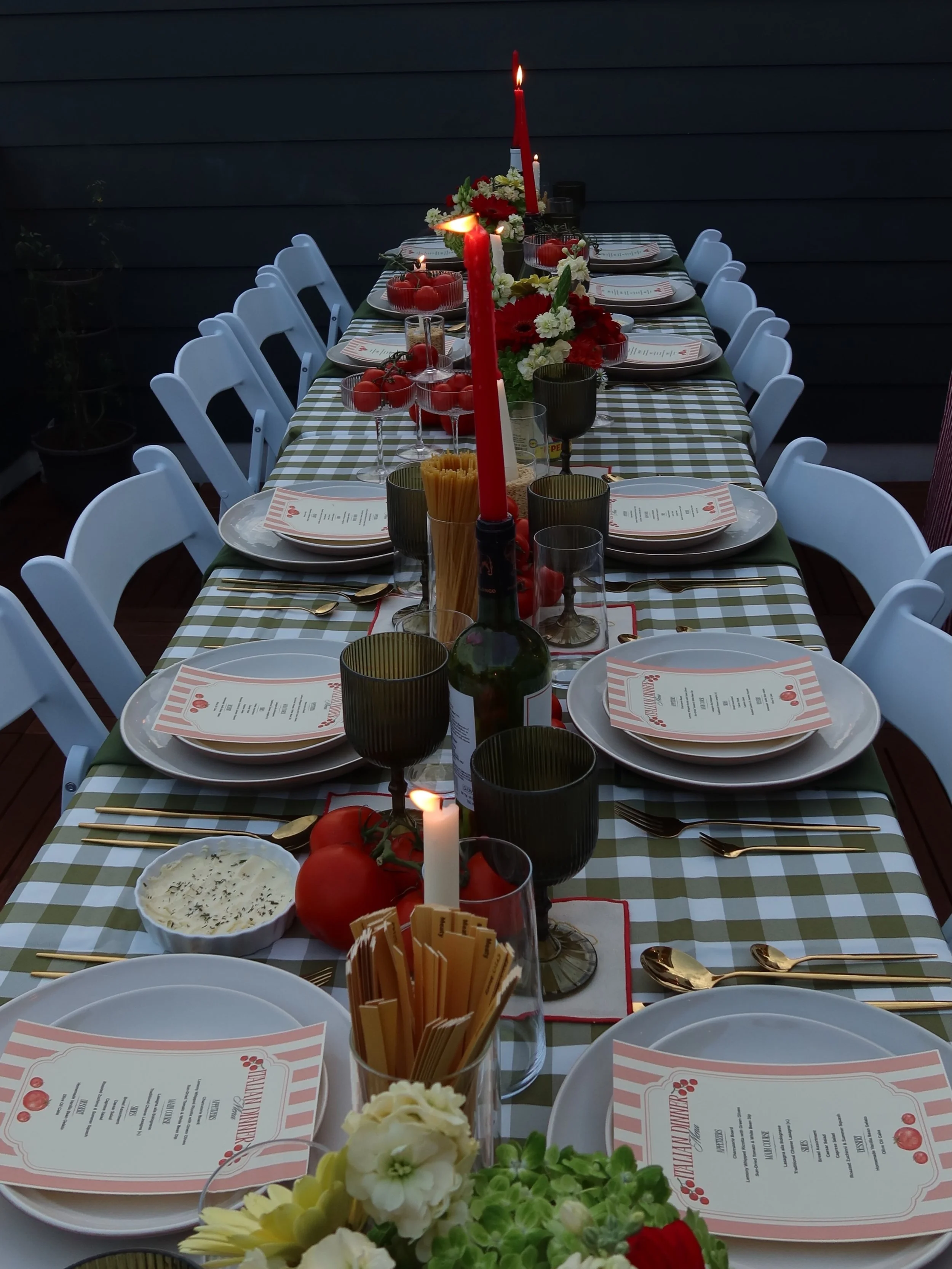 A long outdoor dining table decorated for a celebration with red candles, fresh tomatoes, flowers, and place settings with menus, surrounded by white chairs.