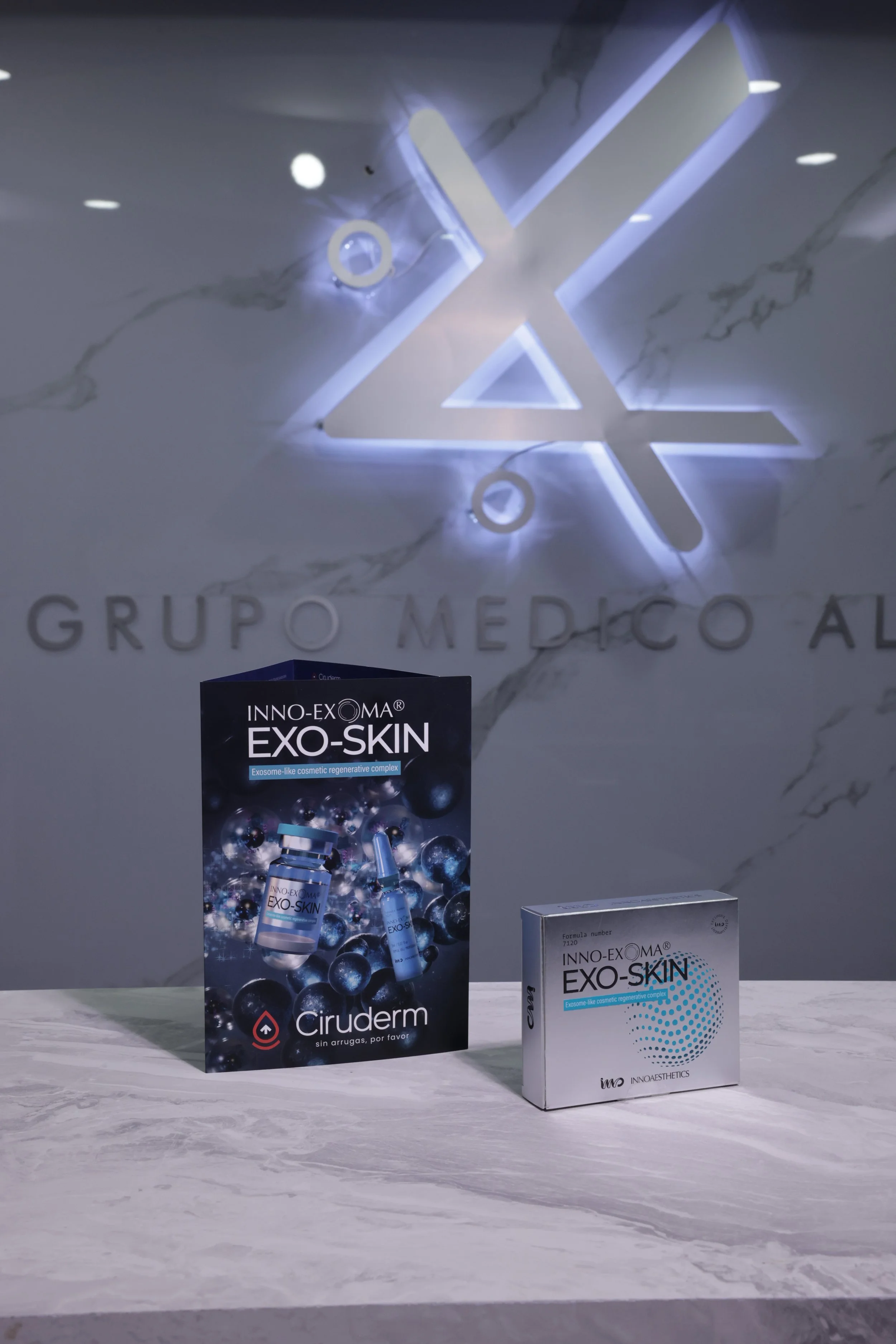 Display of skincare products, including a box and a brochure for INNO-EXOMA EXO-SKIN, on a marble surface in front of a wall with a logo and the words 'GRUPO MÉDICO AL' illuminated.