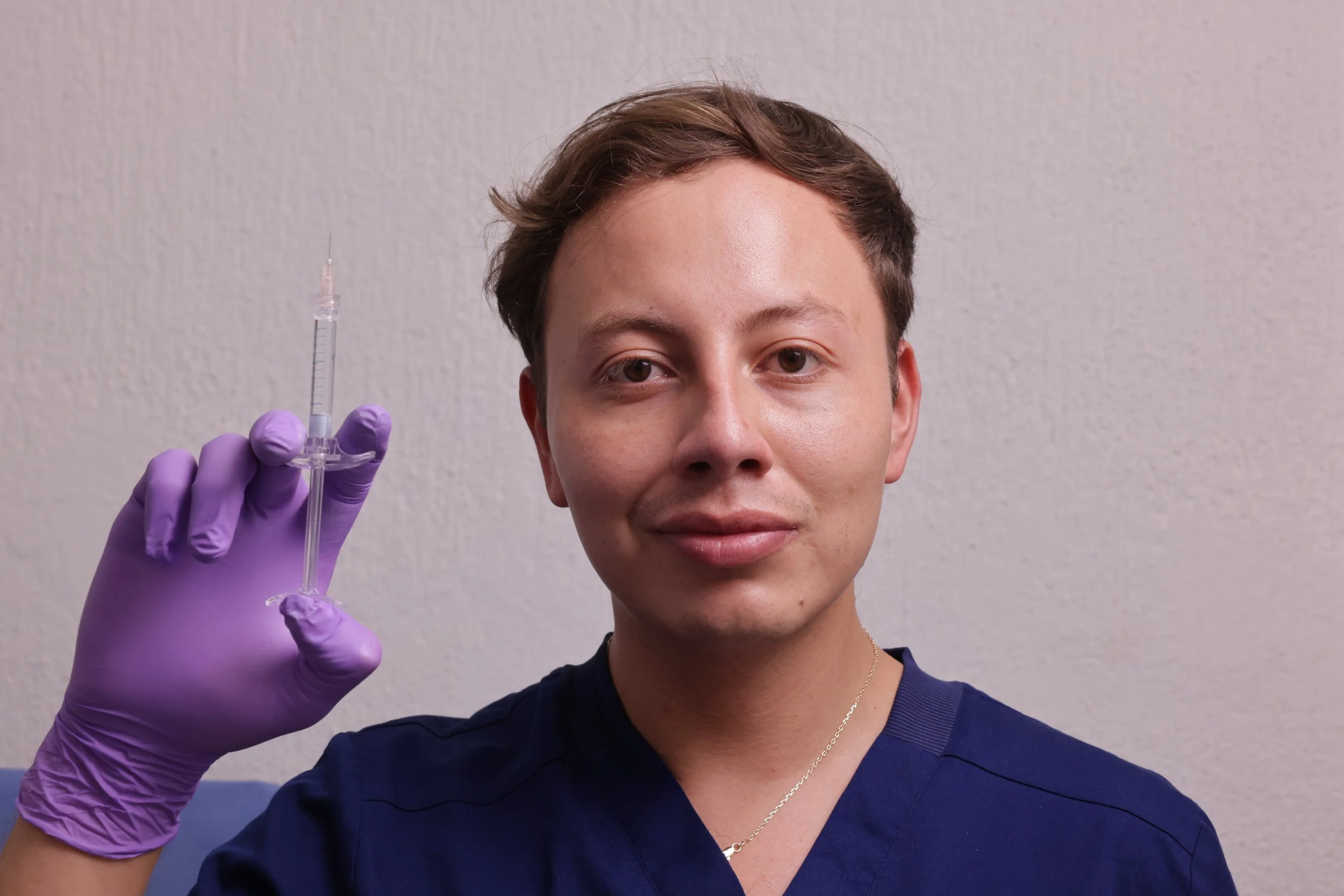 Young woman with short brown hair holding a syringe in her gloved hand. She is wearing a dark shirt and silver necklace, standing against a plain wall background.